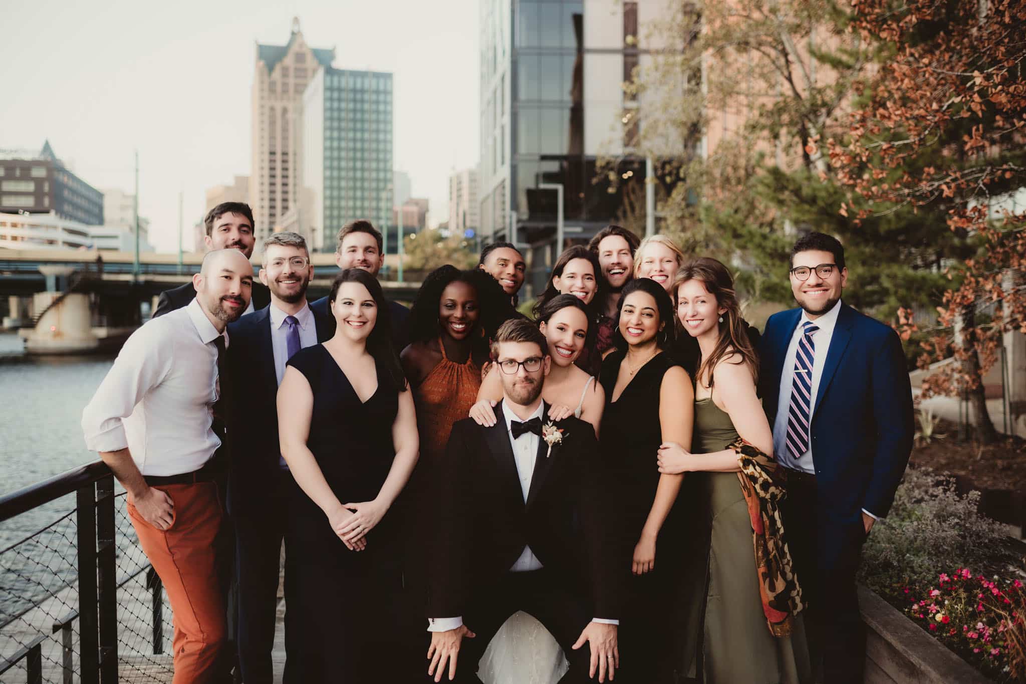 diverse_wedding_party_urban_setting A diverse group of people in formal attire celebrating at a wedding, standing on a city bridge with modern buildings in the background