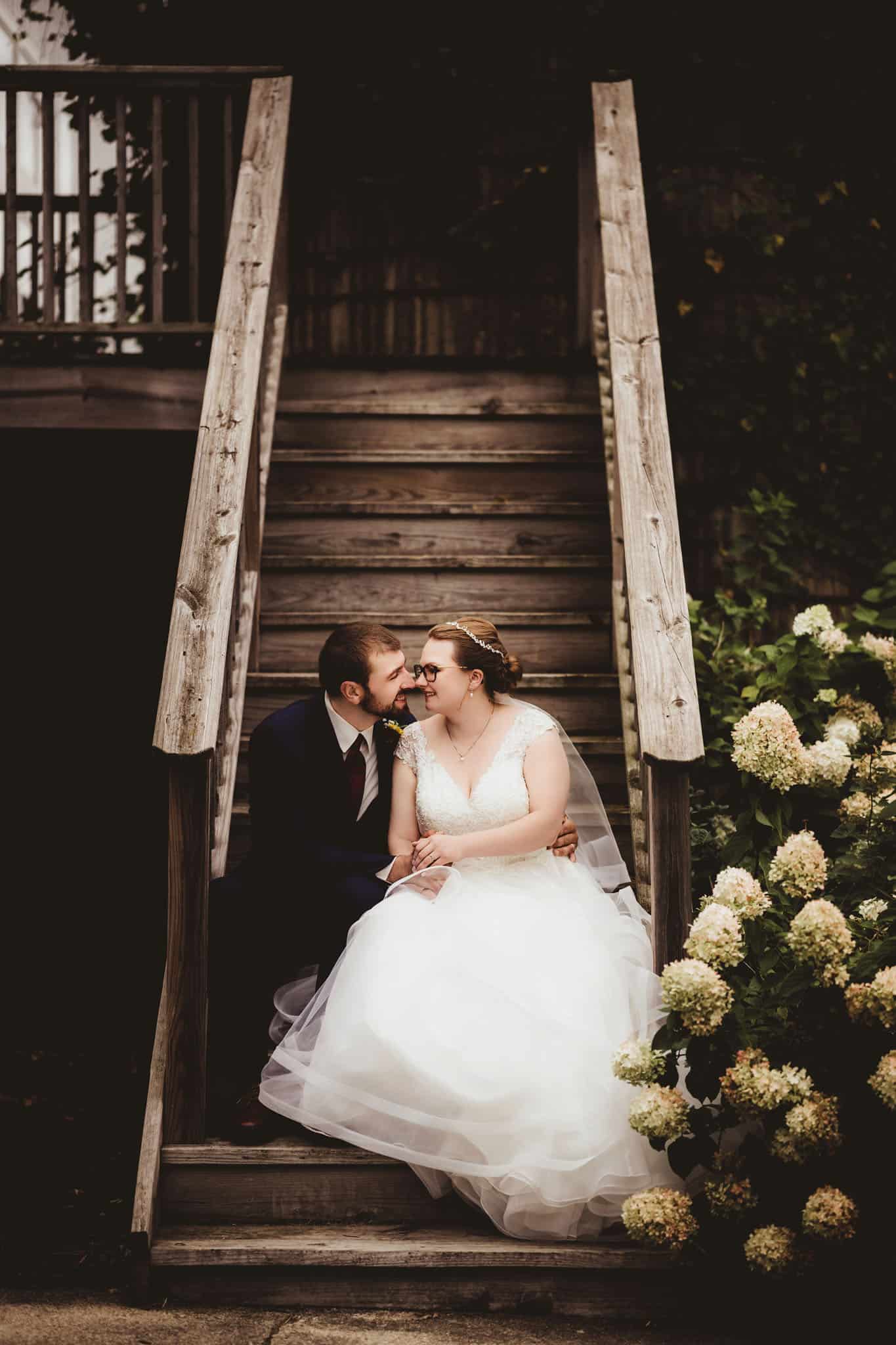 romantic_wedding_couple_on_wooden_stairs A bride in a white dress and a groom in a navy suit seated closely together on aged wooden stairs, surrounded by lush greenery.