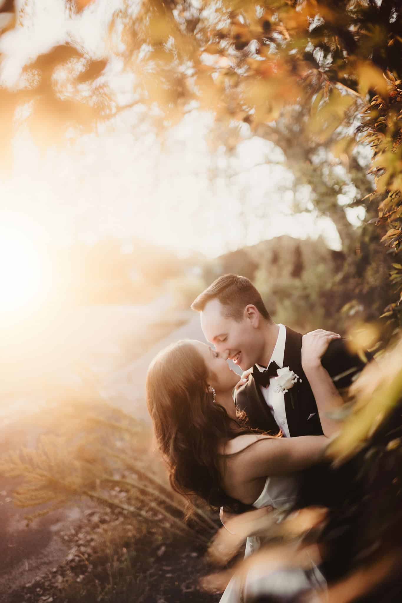 romantic_wedding_couple_sunset_embrace A wedding couple embracing joyfully in a sunlit, autumnal setting.
