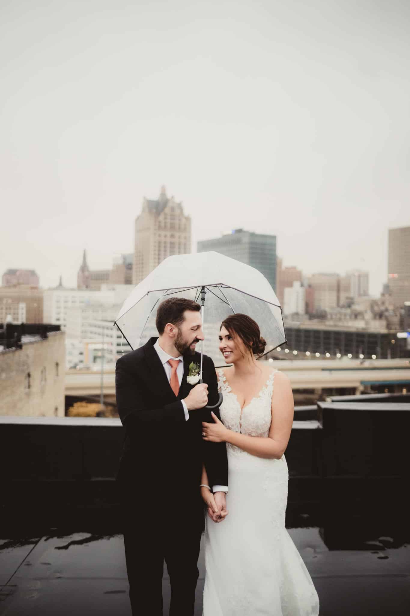urban_rooftop_wedding_couple_with_umbrella A newlywed couple sharing a romantic moment under a clear umbrella on a city rooftop, with skyline in the background.