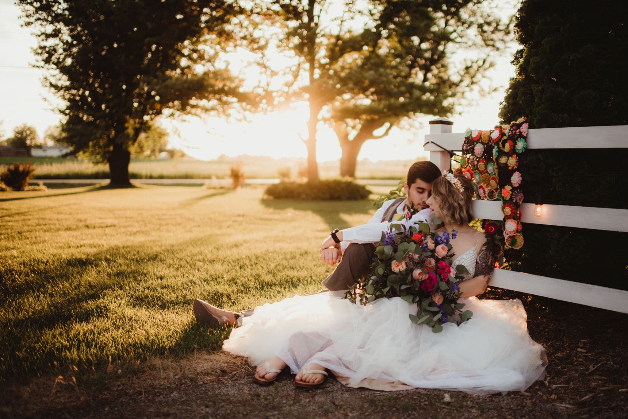 romantic_wedding_couple_sunset_garden A romantic scene of a wedding couple seated by a white fence adorned with flowers at sunset, with a large tree and soft golden light in the background.