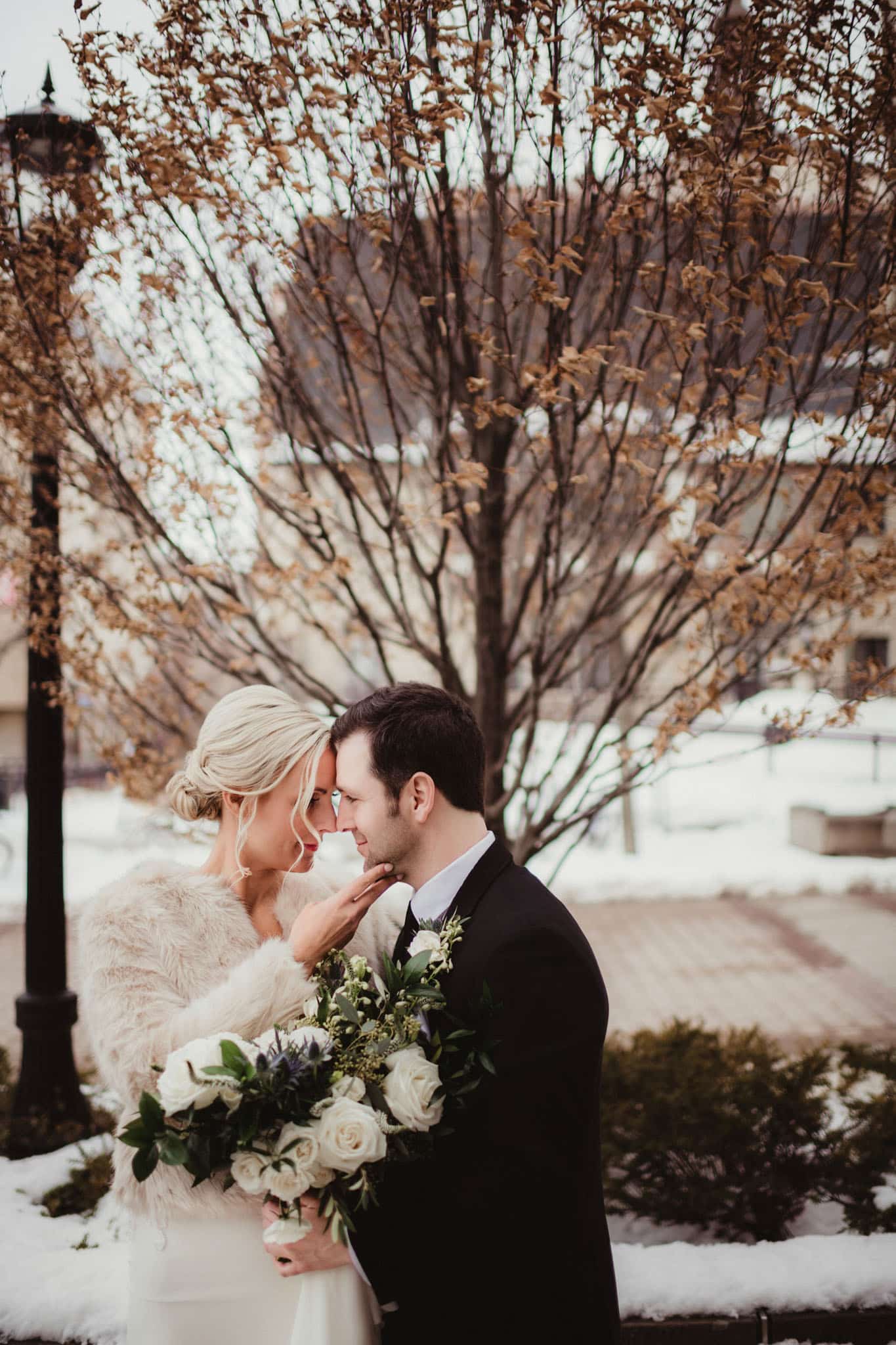 winter_wedding_couple_embrace_outdoors Bride and groom sharing a tender moment in a snowy setting, surrounded by bare trees and white flowers