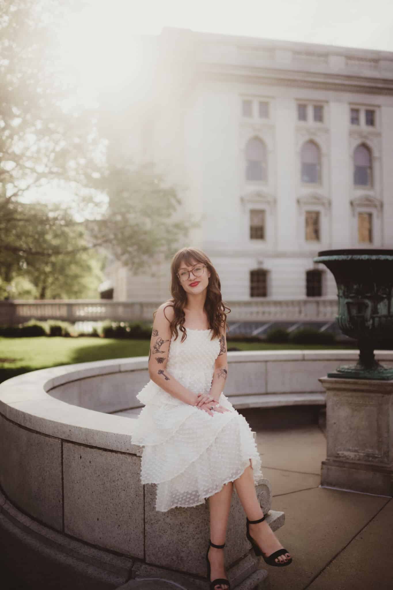 woman_in_white_dress_sitting_at_courthouse A young woman with tattoos wearing a white dress and glasses, sitting on the edge of a fountain with a large courthouse in the background during sunset