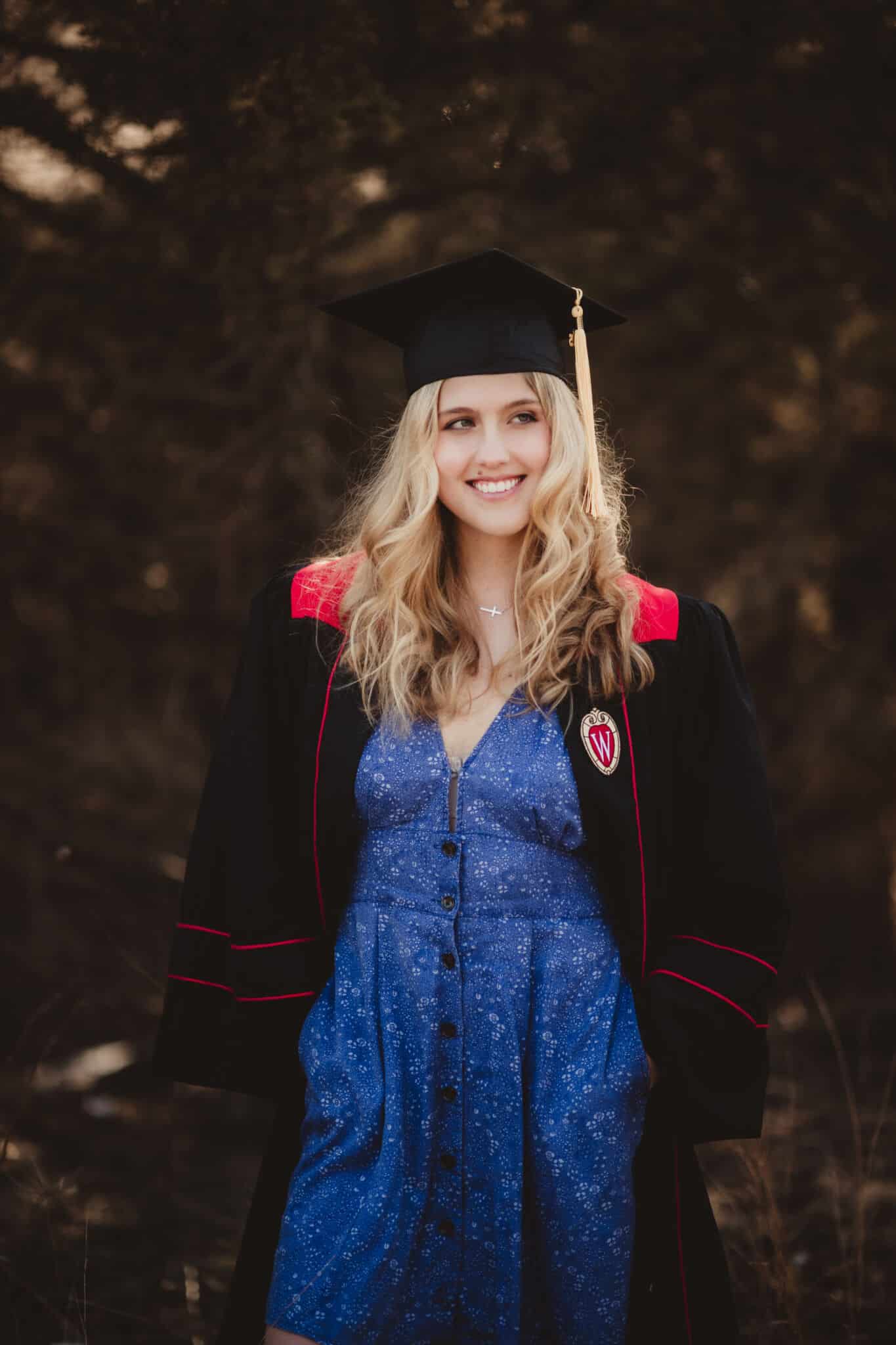 smiling_female_graduate_in_wisconsin_university_regalia Young woman with a radiant smile, dressed in Wisconsin University graduation regalia, standing in a wooded area