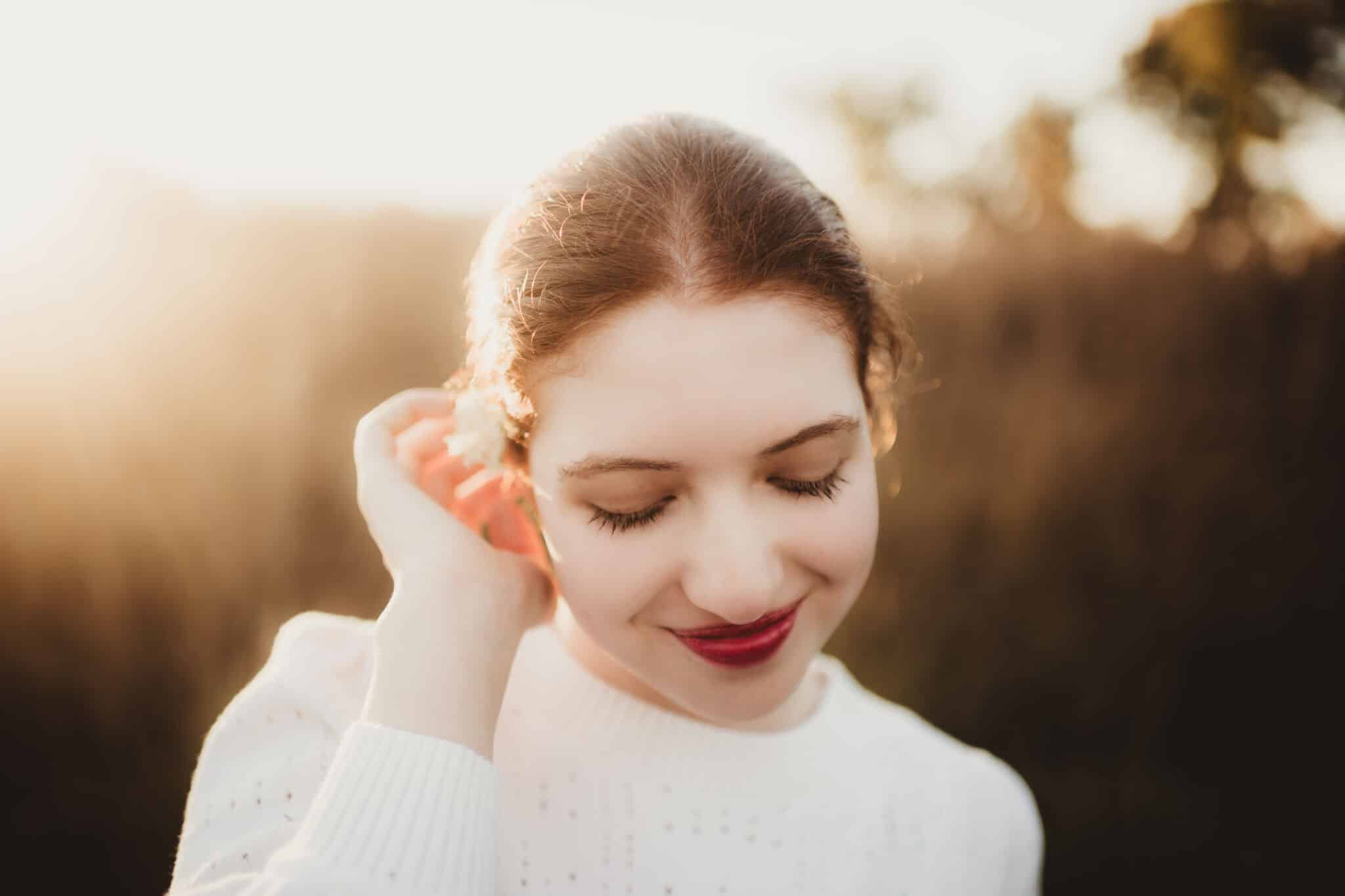 young_woman_enjoying_sunlight_in_field Young woman with red hair smiling gently, holding a flower, bathed in golden sunlight with a blurred natural background