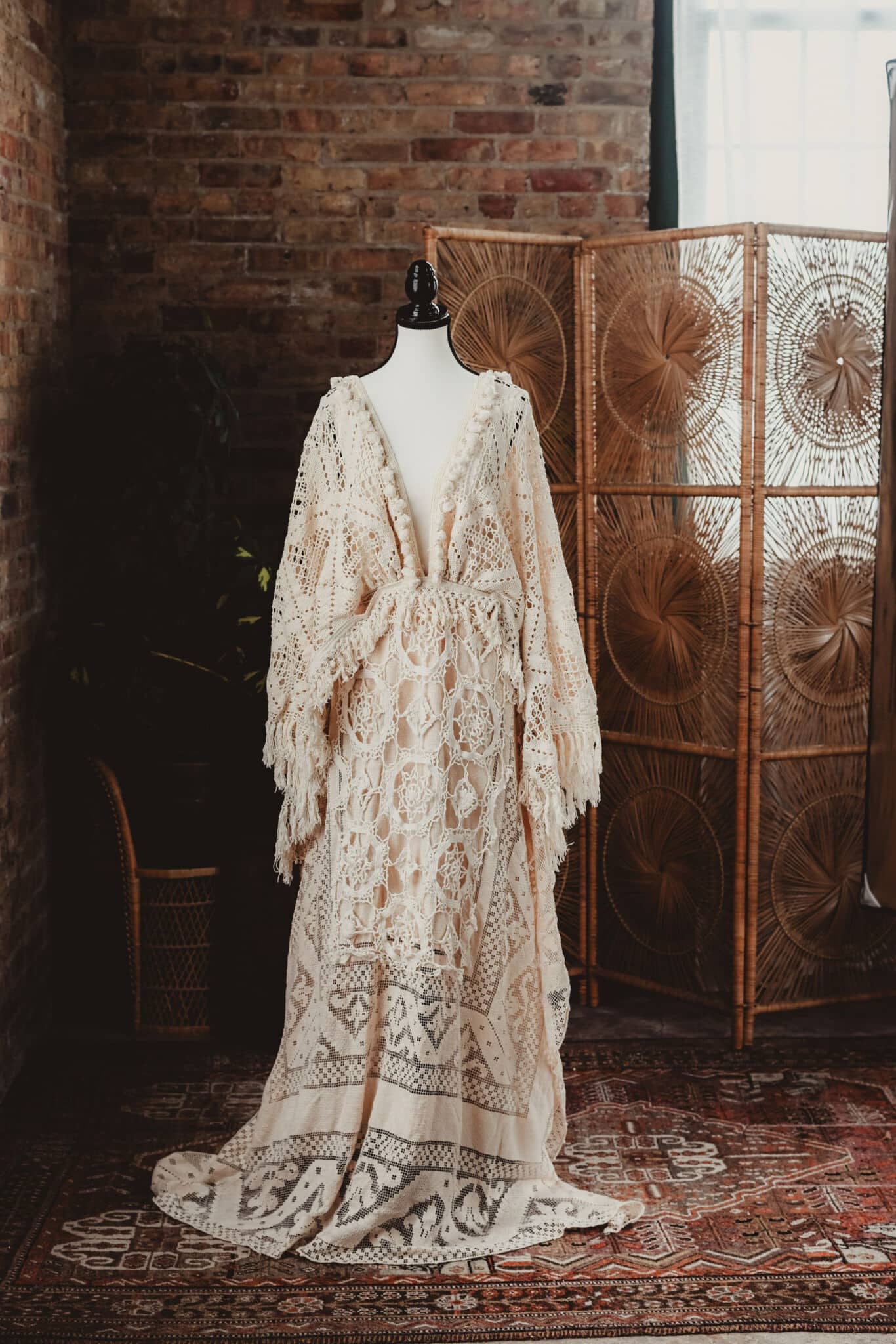 An elegant vintage lace dress displayed on a mannequin in front of a brick wall with a woven room divider in the background.