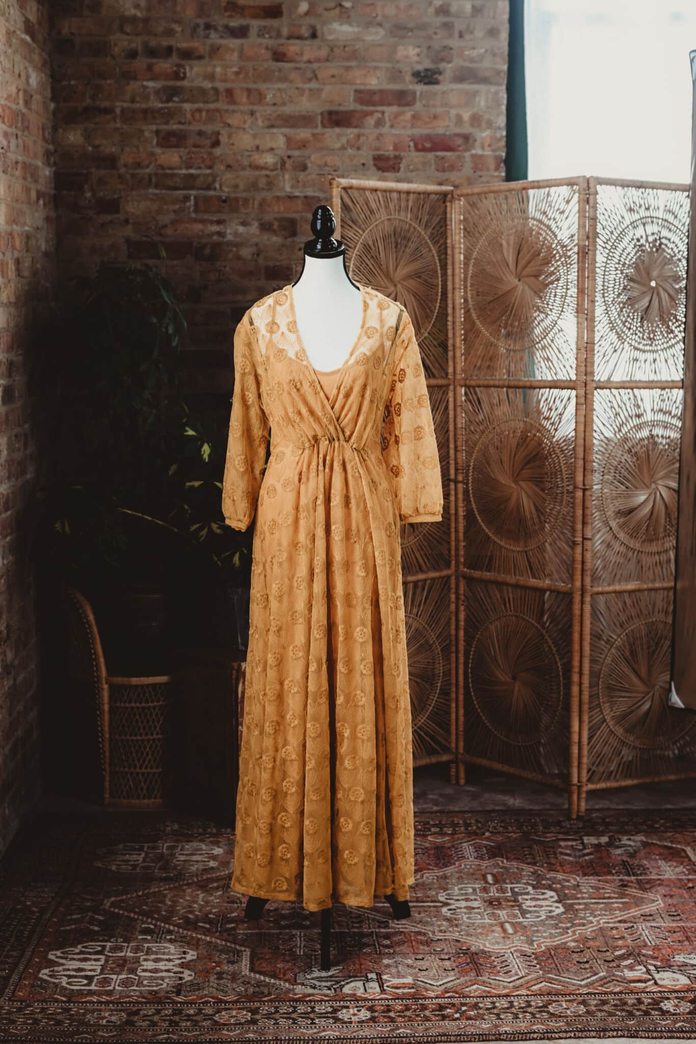 A vintage yellow dress displayed on a mannequin in front of a woven screen and a brick wall, standing on an oriental rug.