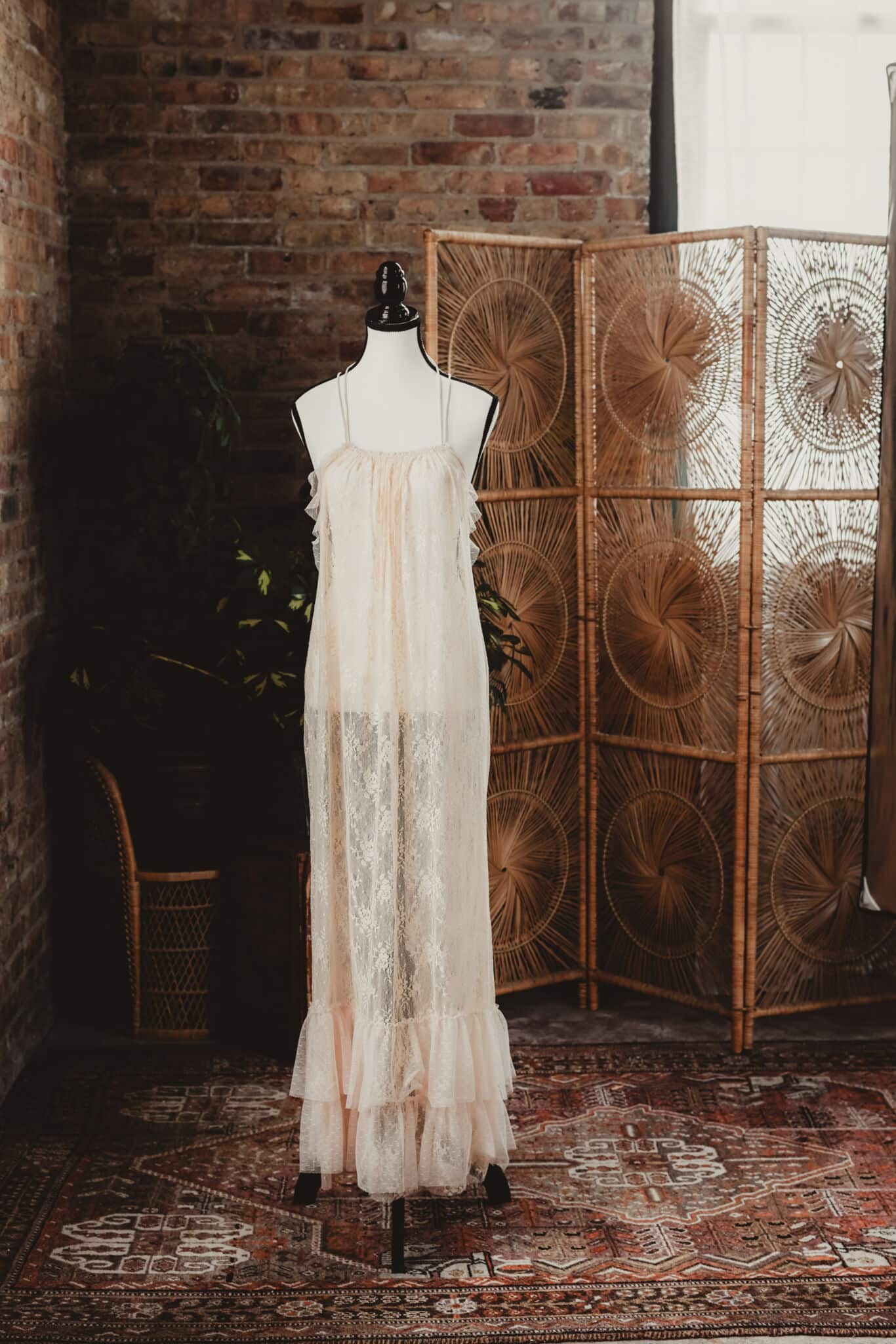 An elegant vintage lace dress displayed on a mannequin against a rustic brick wall backdrop, with intricate wicker dividers and lush greenery in the background.