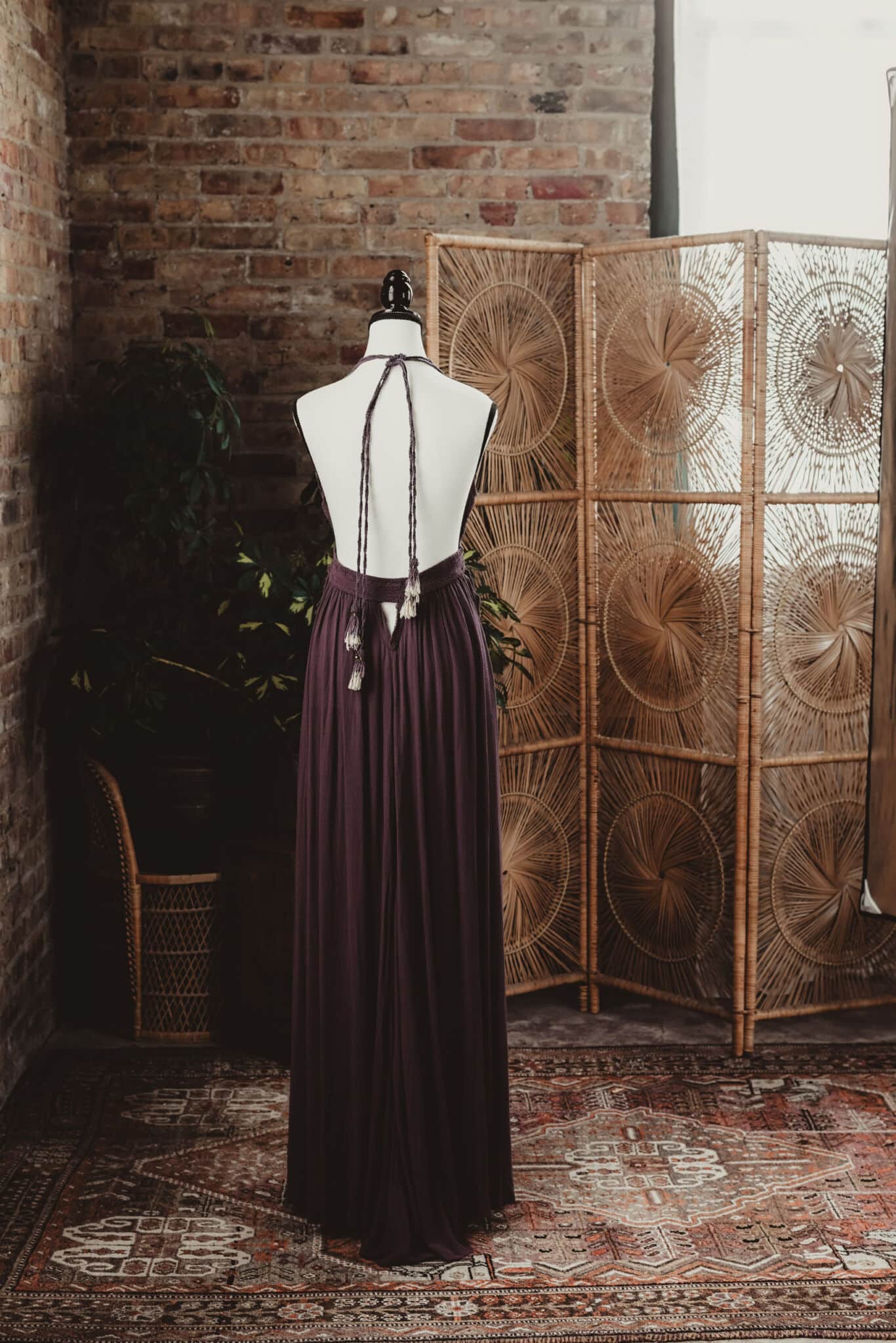 A mannequin dressed in a long, burgundy bohemian gown set against a rustic brick wall with a folding rattan room divider in the background.