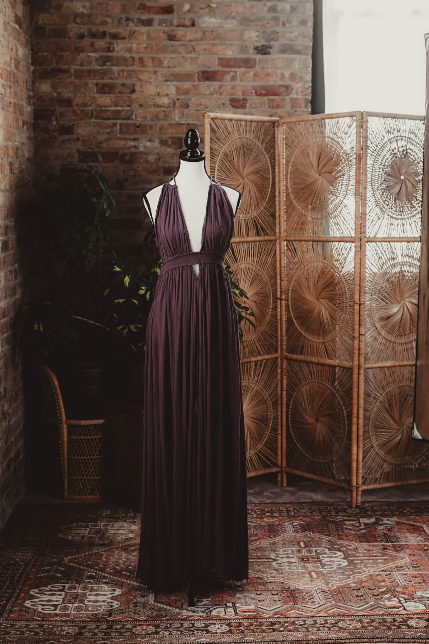 A mannequin draped in a flowing purple gown posed against a backdrop of a rustic brick wall and decorative rattan room divider