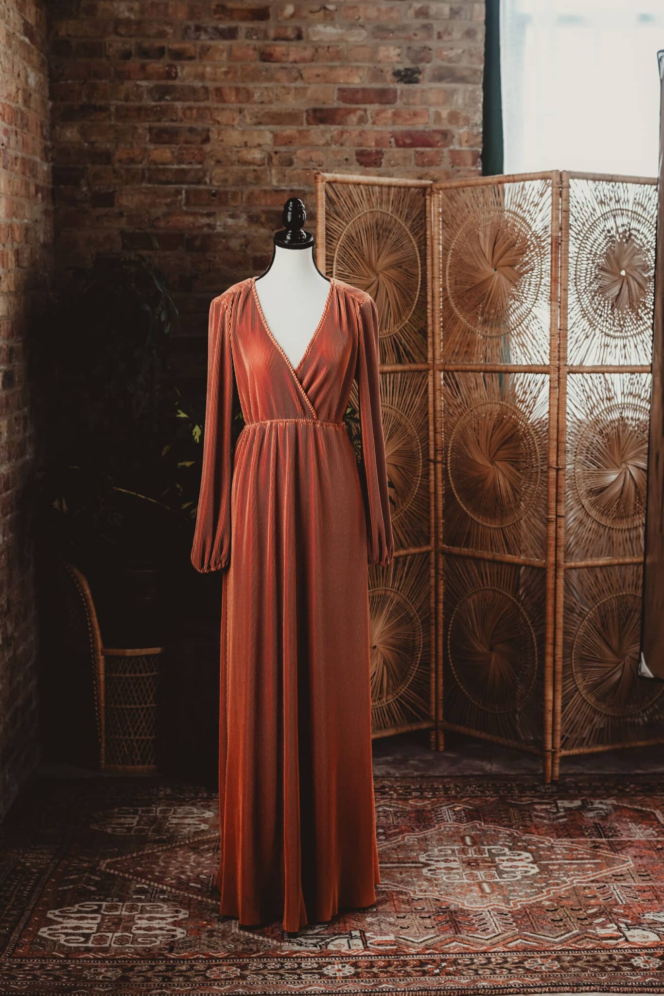 An elegant vintage orange dress displayed on a mannequin in a rustic room with a brick wall and a decorative wicker screen.