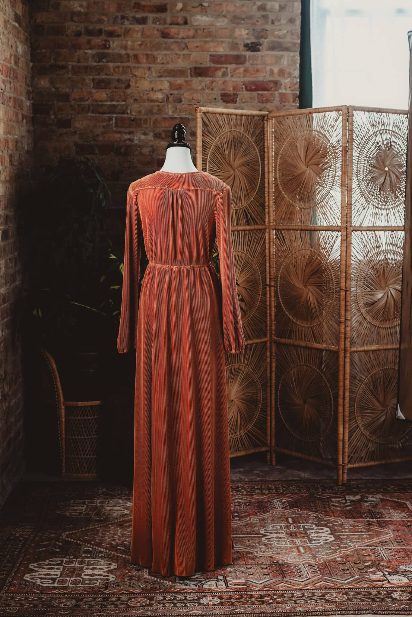A vintage orange pleated dress on a mannequin against a brick wall backdrop with a decorative wicker room divider and lush plants.