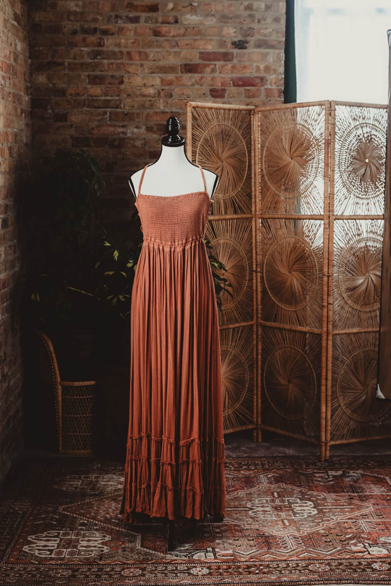 A rust-colored vintage dress on a mannequin against an exposed brick wall backdrop, complemented by a wicker room divider