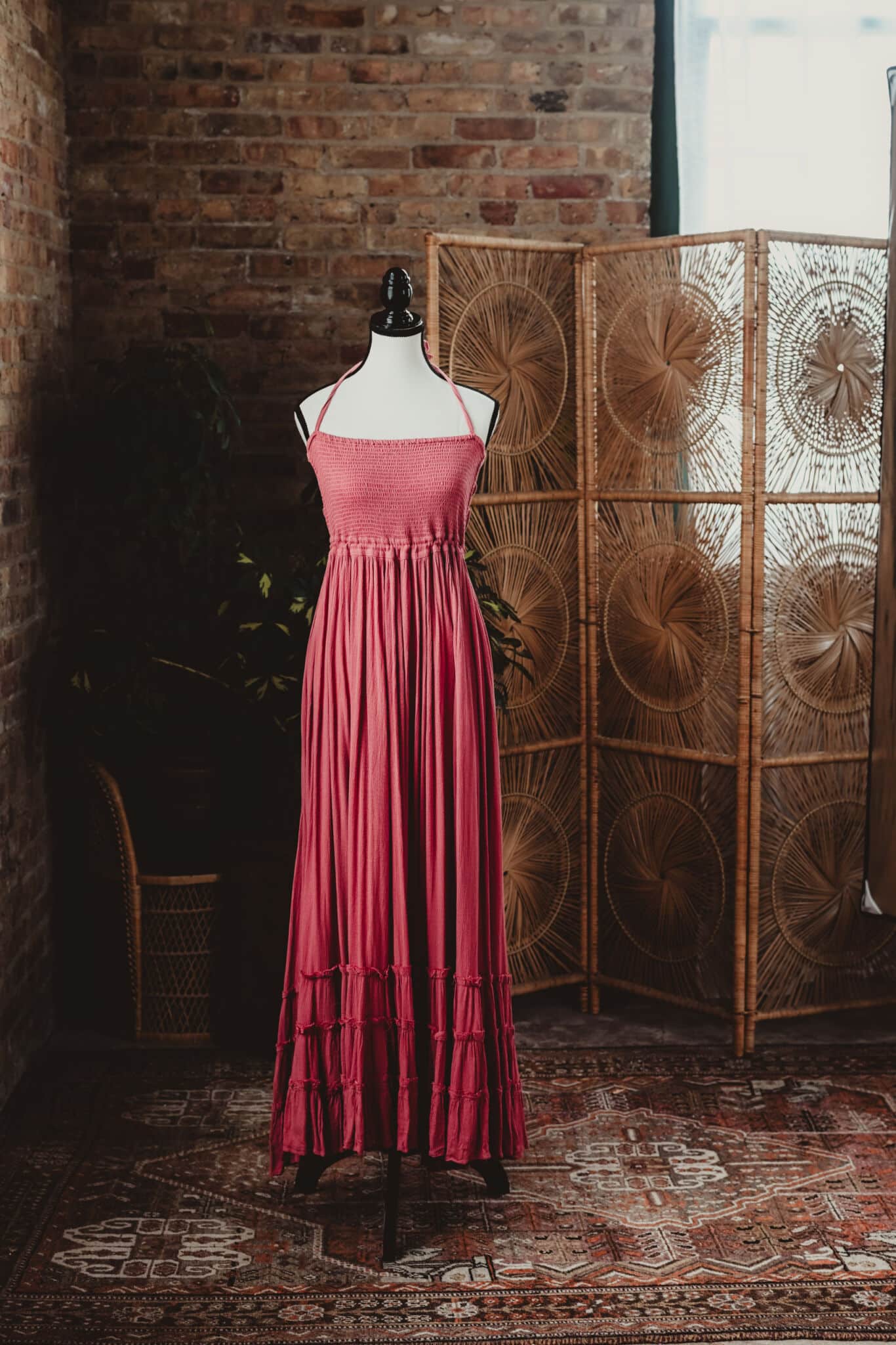 Pink summer dress displayed on a black mannequin against a rustic brick wall and woven room divider