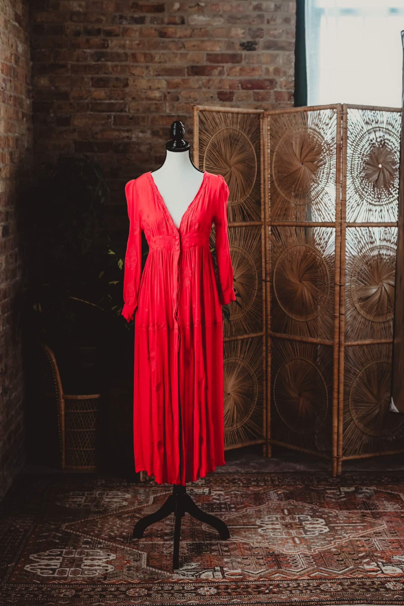 A vibrant red vintage dress displayed on a mannequin against a rustic brick wall backdrop with a decorative wicker screen.