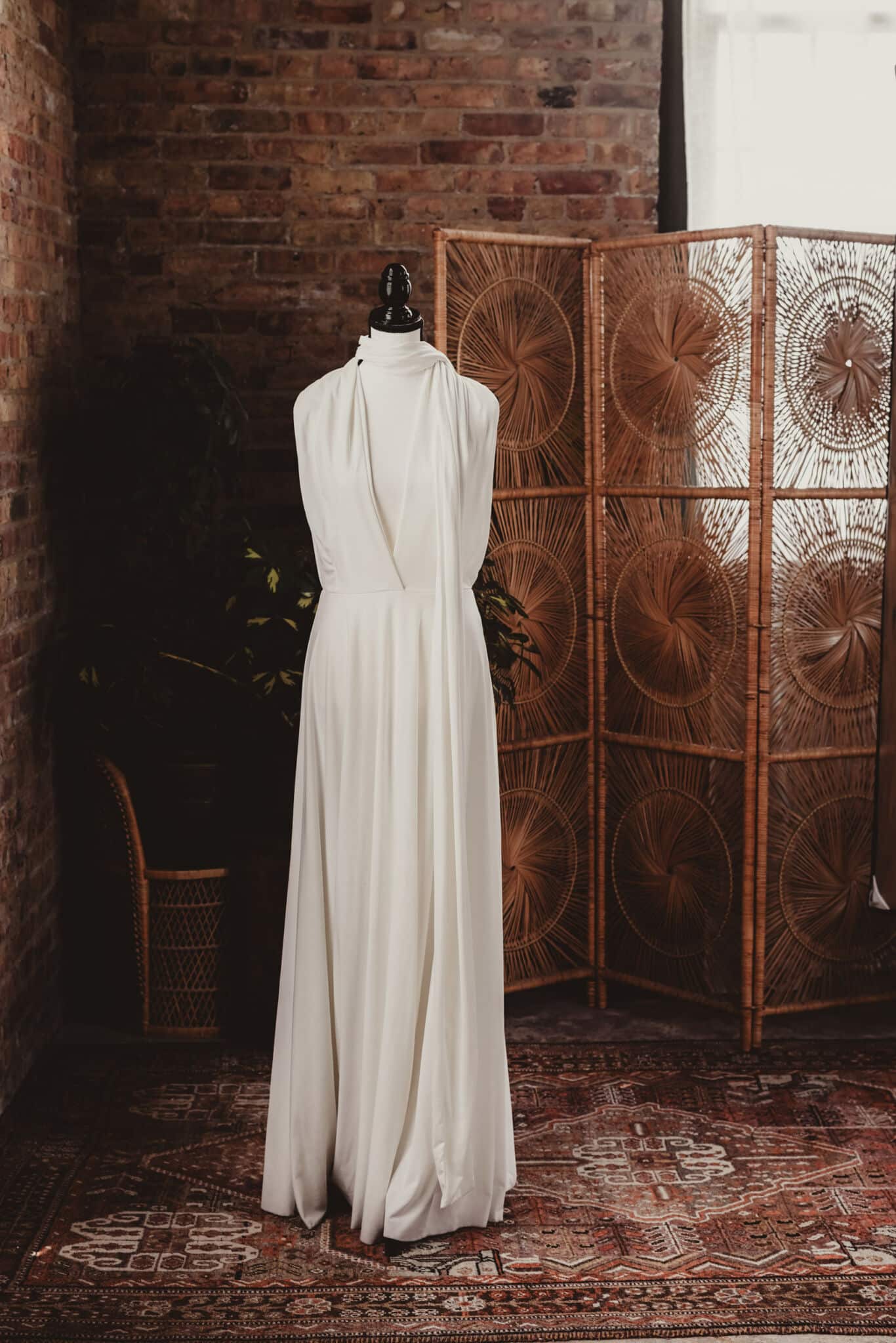 An elegant white dress displayed on a mannequin in a rustic room with a brick wall and a decorative woven screen backdrop.