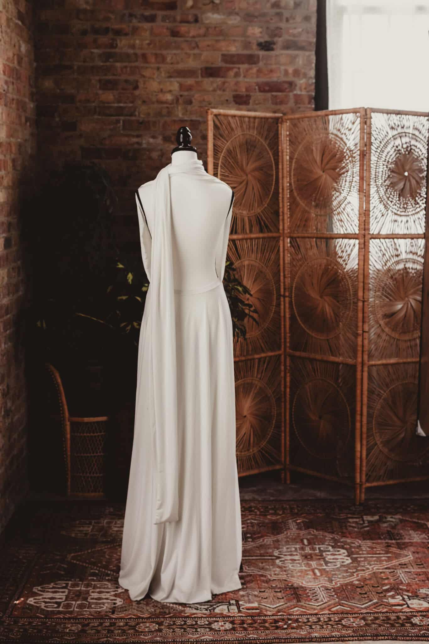 An elegant white long-sleeved dress draped on a mannequin, positioned against an intricate brick wall backdrop with a bohemian rattan screen and exotic plants.