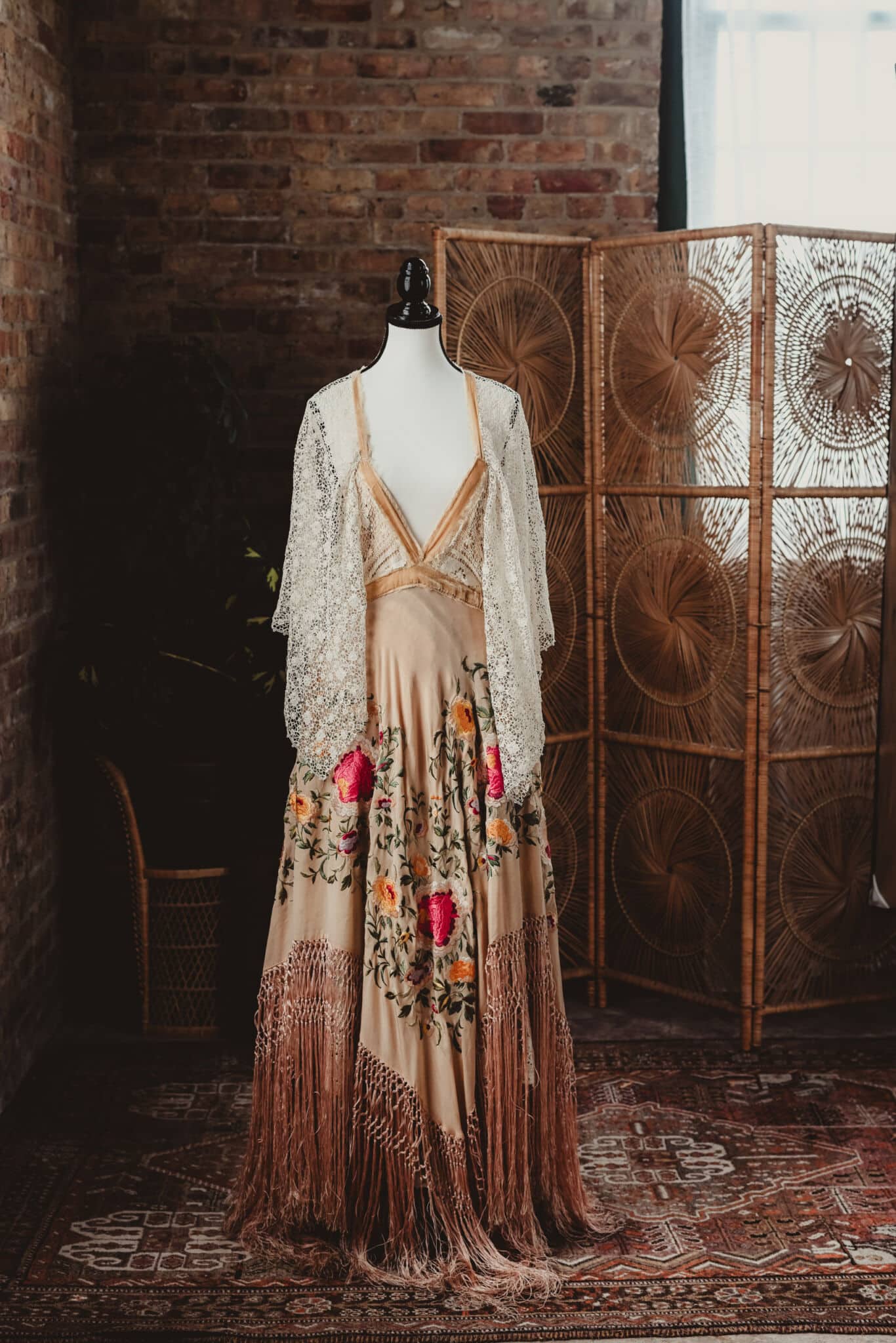 An ornately embroidered vintage gown on a mannequin, featuring vibrant floral designs and a lace shawl, against a rustic brick background with a wicker screen.