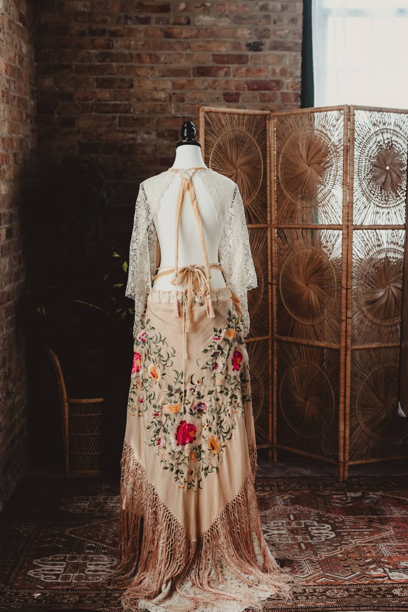 A vintage, embroidered dress displayed on a mannequin, set against a bohemian-style room backdrop with woven decorations and a brick wall.