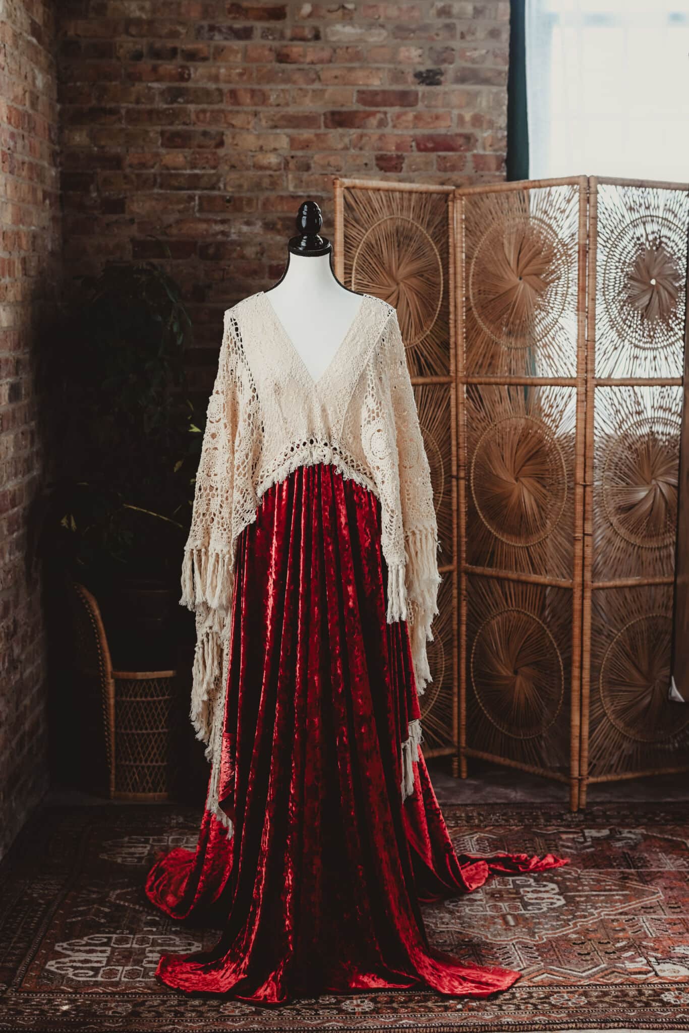 Elegant vintage dress with long red velvet skirt and cream crochet top displayed on a mannequin against a rustic brick wall and a woven room divider
