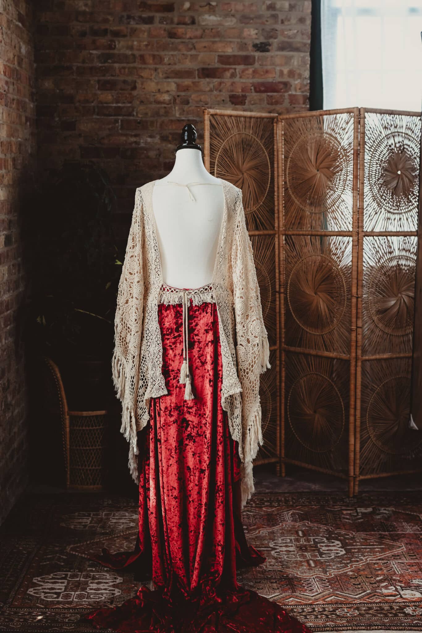 A vintage-inspired mannequin wearing a vibrant red dress paired with a delicate lace shawl, set against a bohemian style woven room divider.