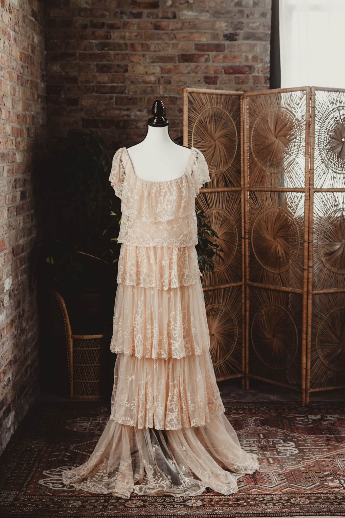 Vintage cream lace wedding dress on a mannequin in a rustic setting with brick wall and bohemian rattan decor