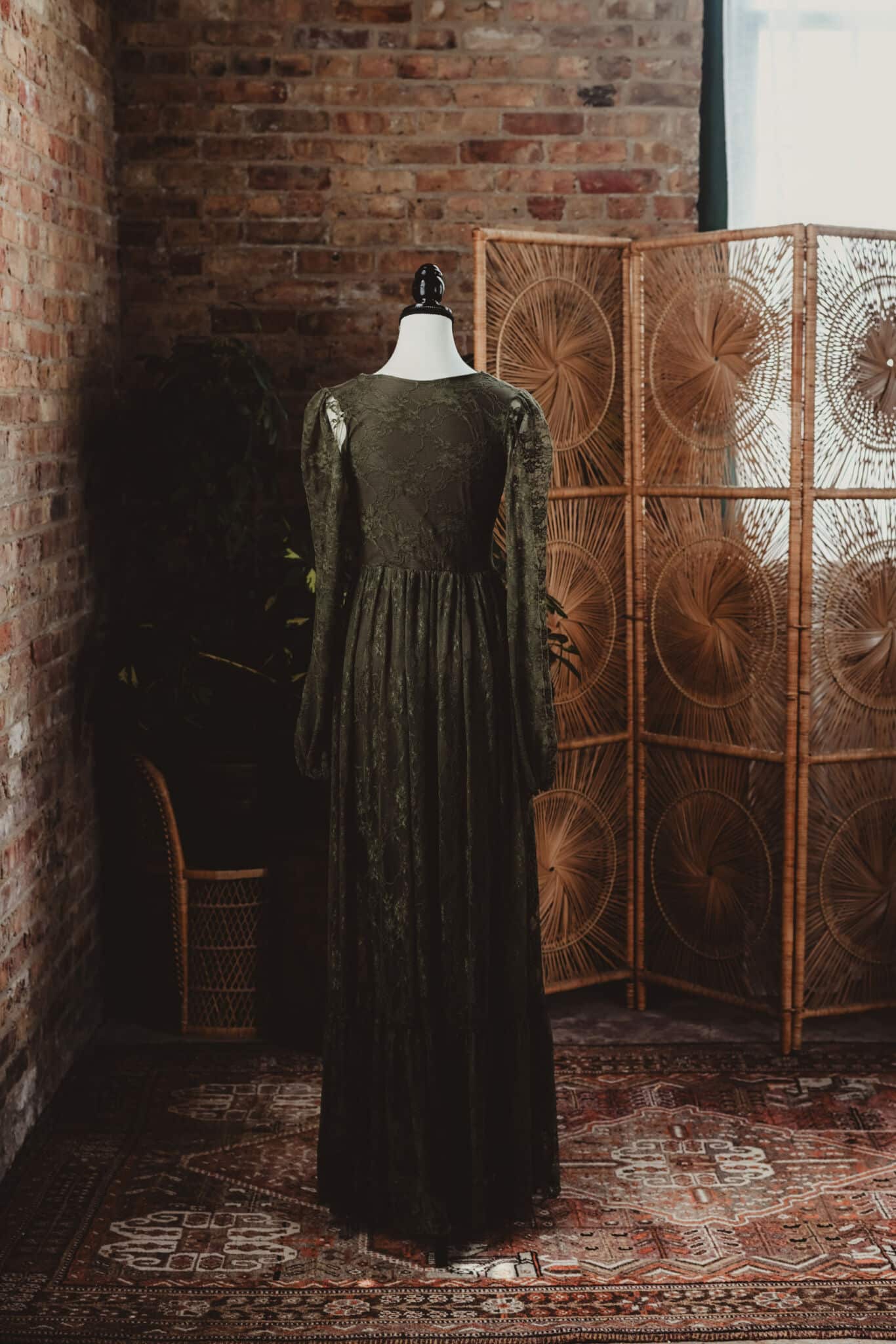 A vintage green lace dress displayed on a mannequin in a rustic room with exposed brick walls and a woven room divider.