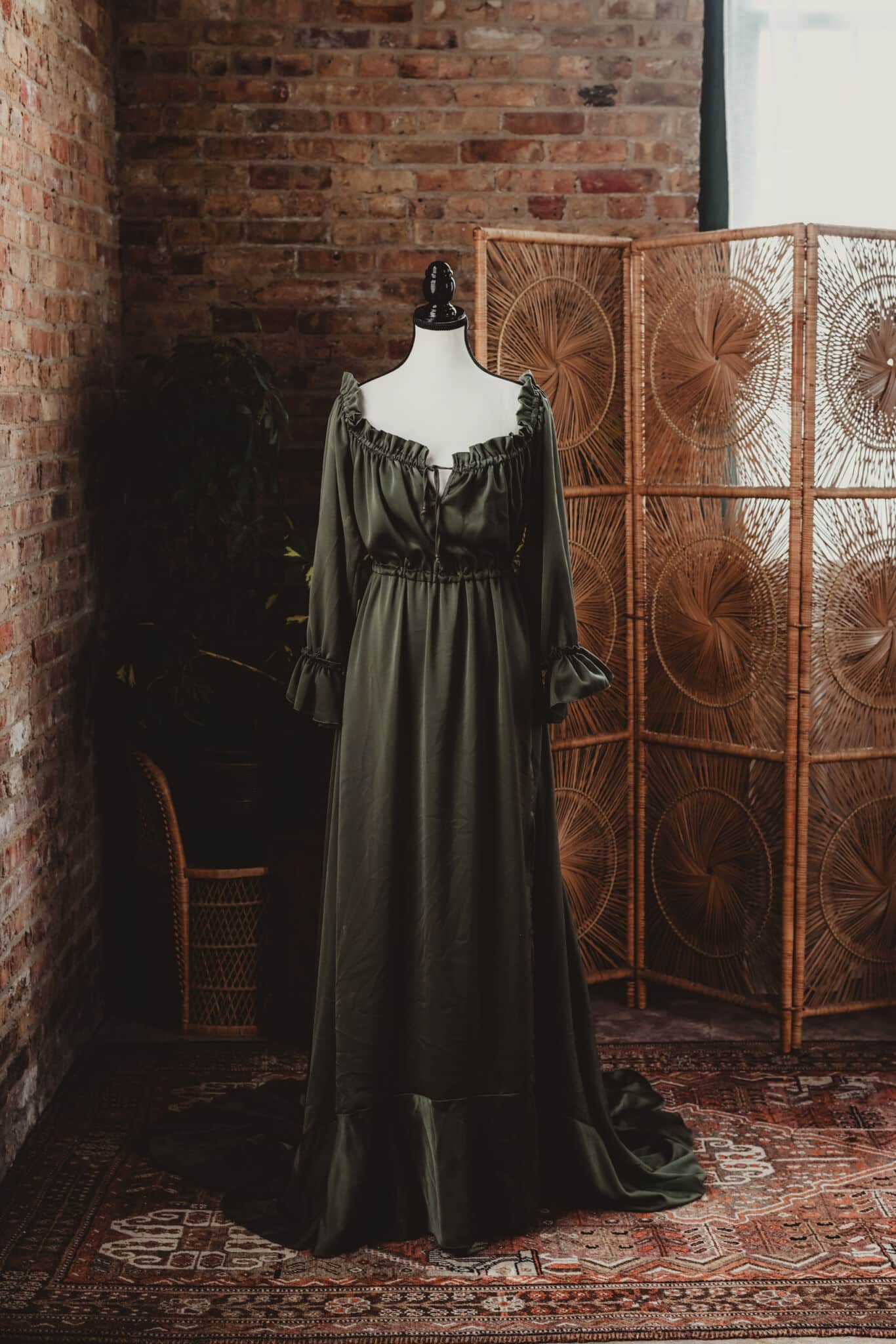 A sophisticated olive green vintage dress displayed on a mannequin in a rustic room with a brick wall and wicker screen backdrop.