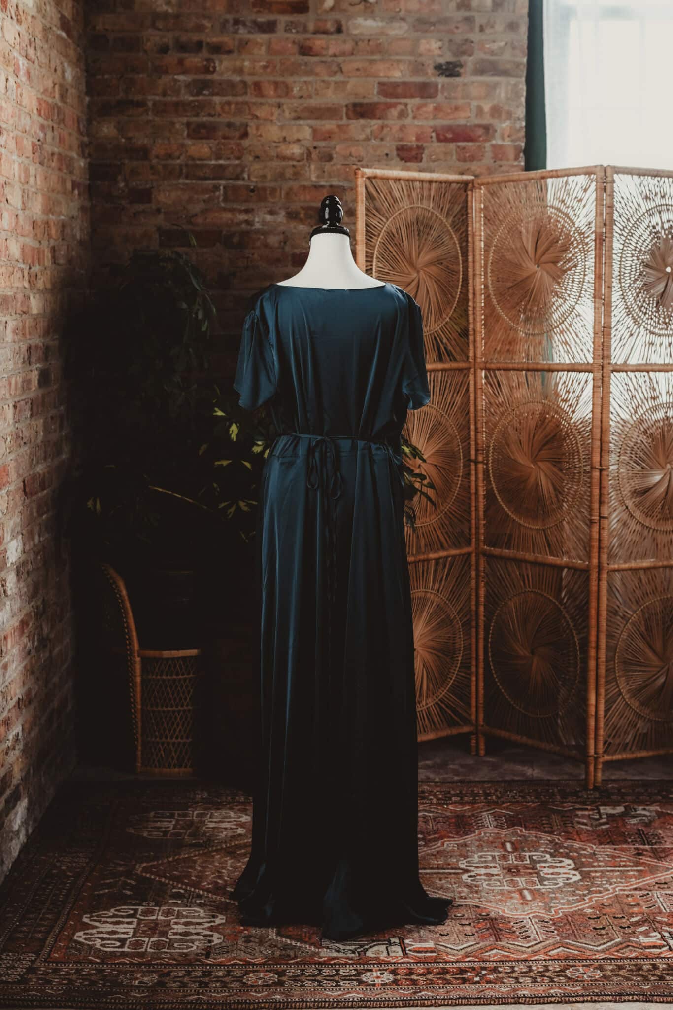A dark blue vintage dress on a mannequin in a rustic room with a brick wall and a wicker room divider