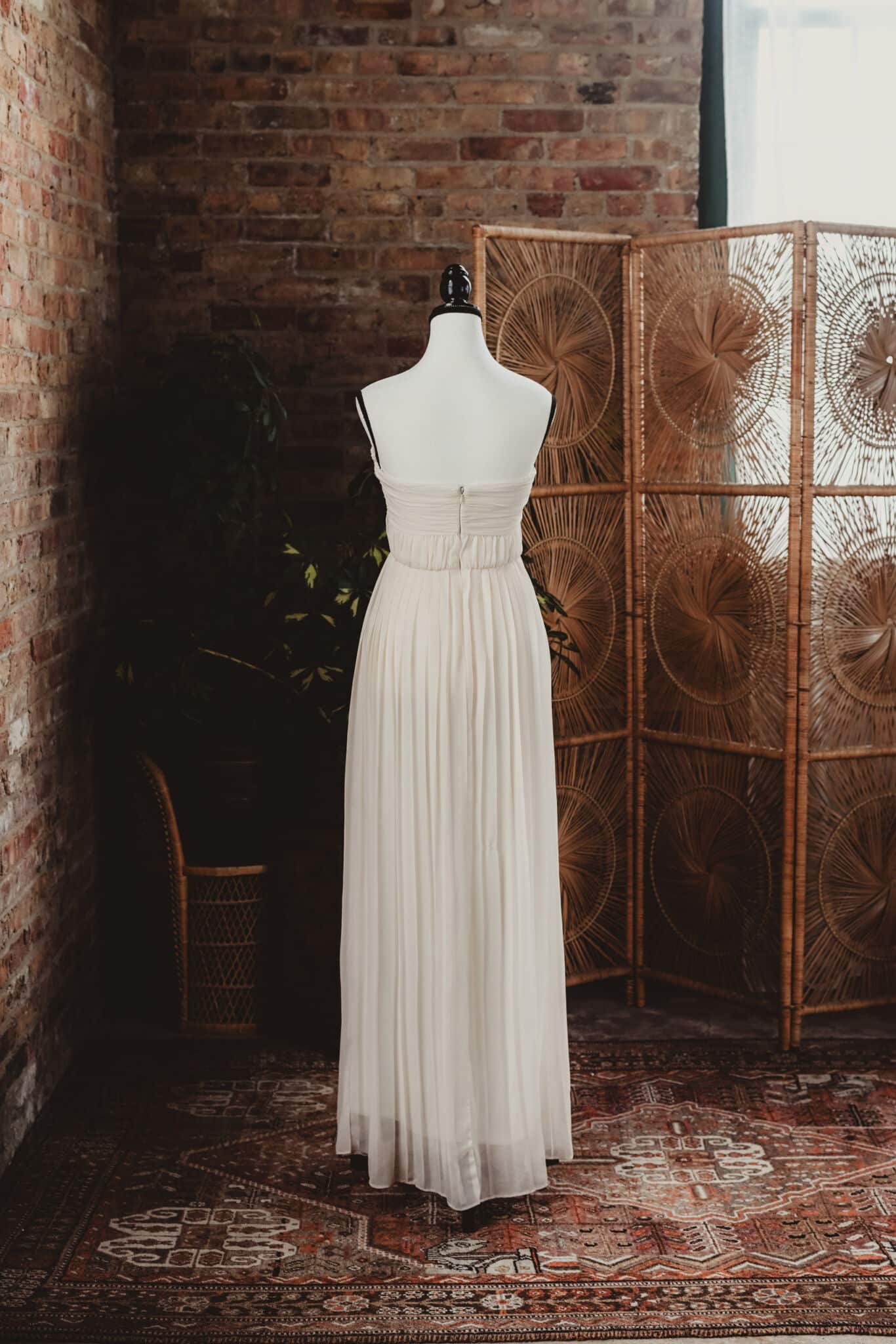 An elegant vintage wedding dress on a mannequin, positioned against a brick wall with a wicker room divider in the background.