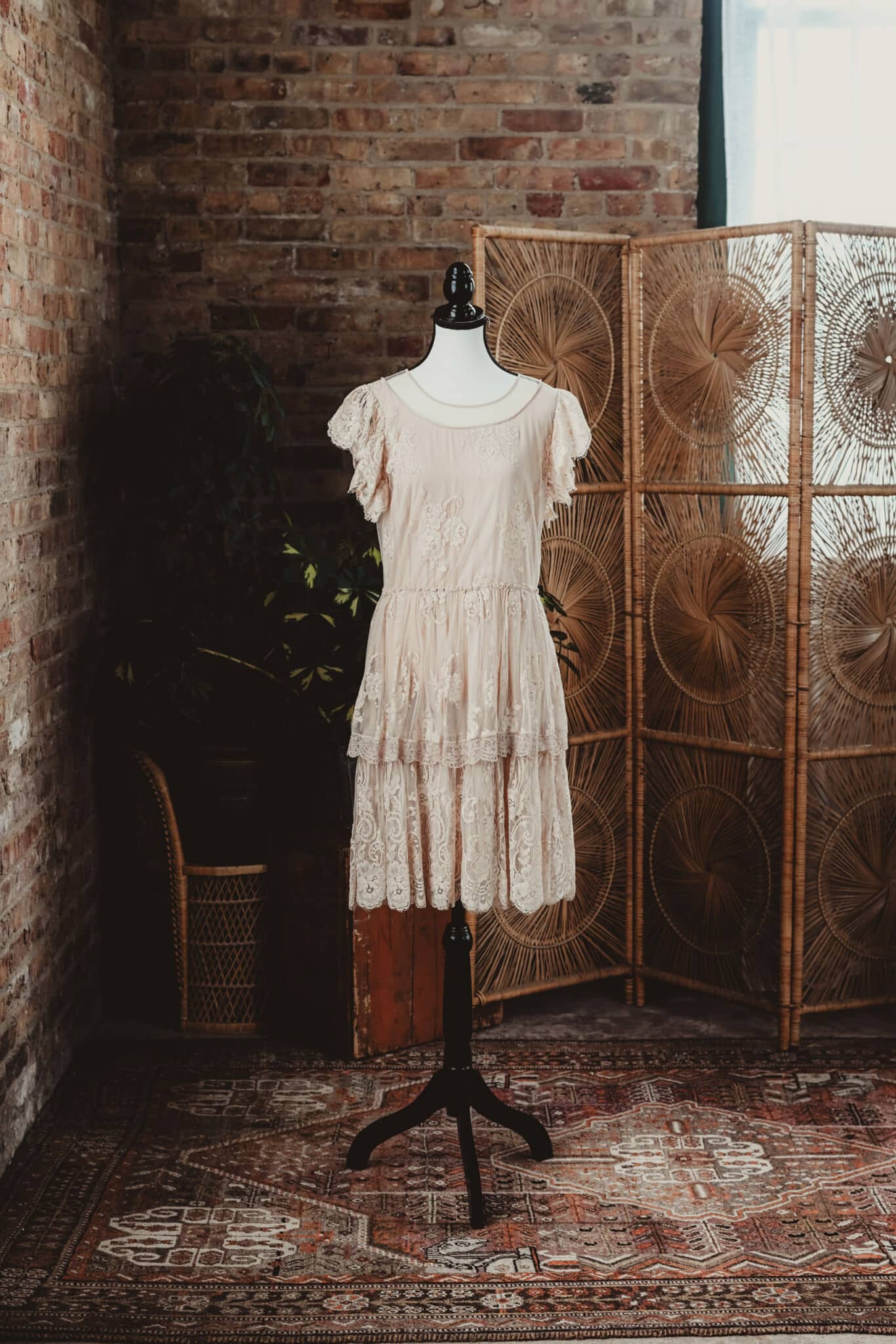 Vintage ivory lace dress displayed on a mannequin in a rustic room with brick walls and a rattan divider.