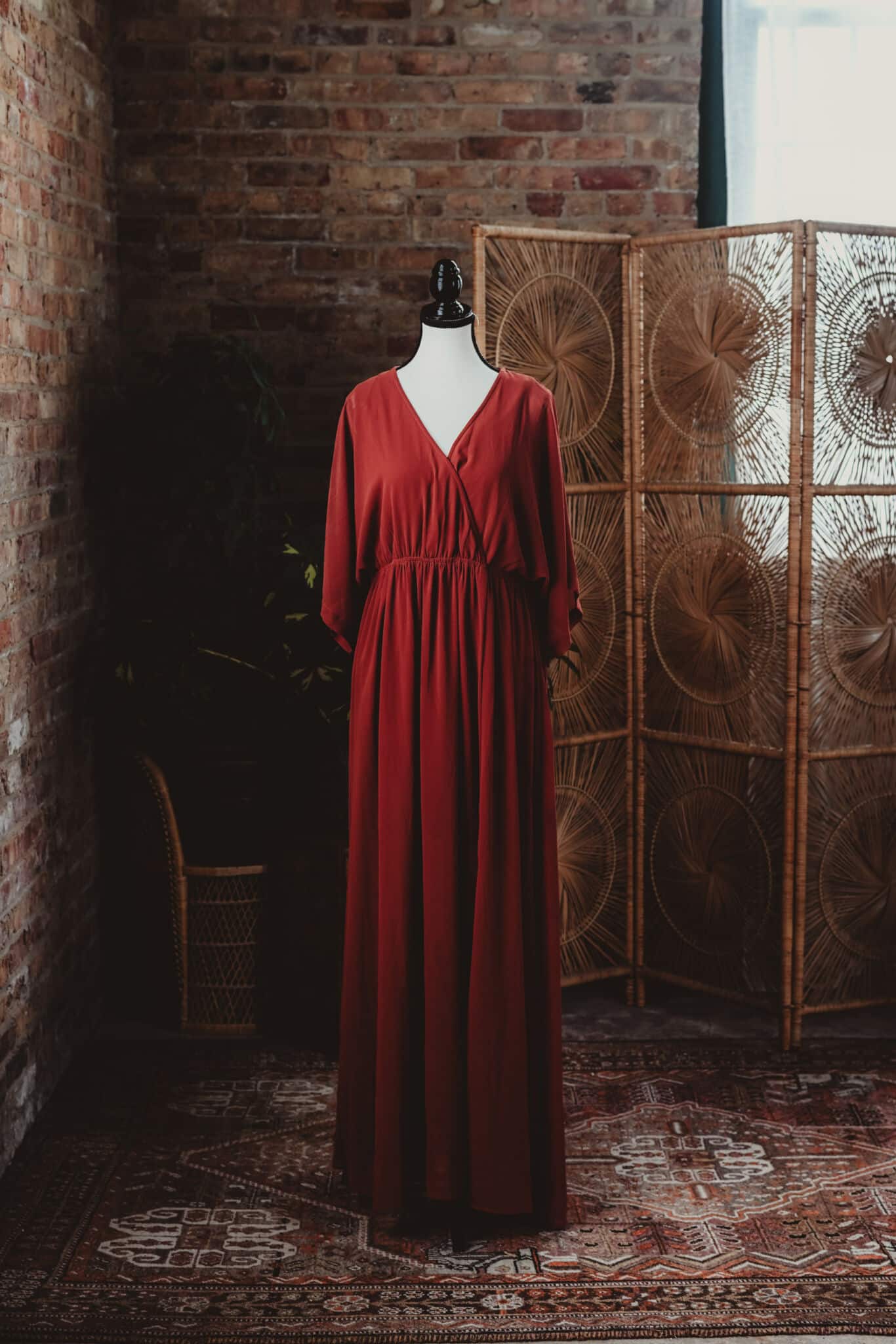 A sophisticated red dress displayed on a mannequin in a rustic room with a brick wall and a decorative wicker screen.