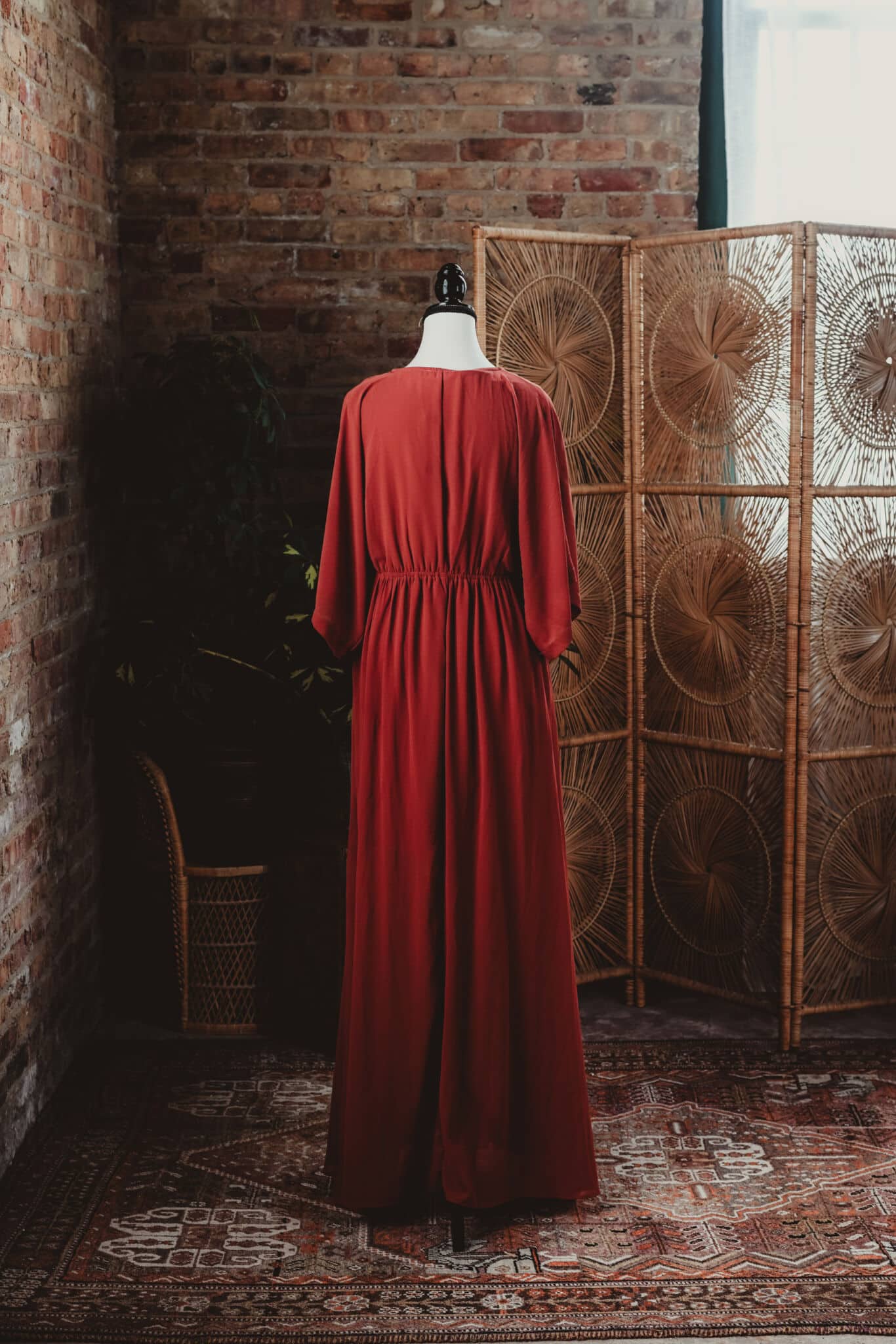 Elegant vintage red dress on a mannequin against a rustic brick wall backdrop with decorative rattan screen and lush indoor plants