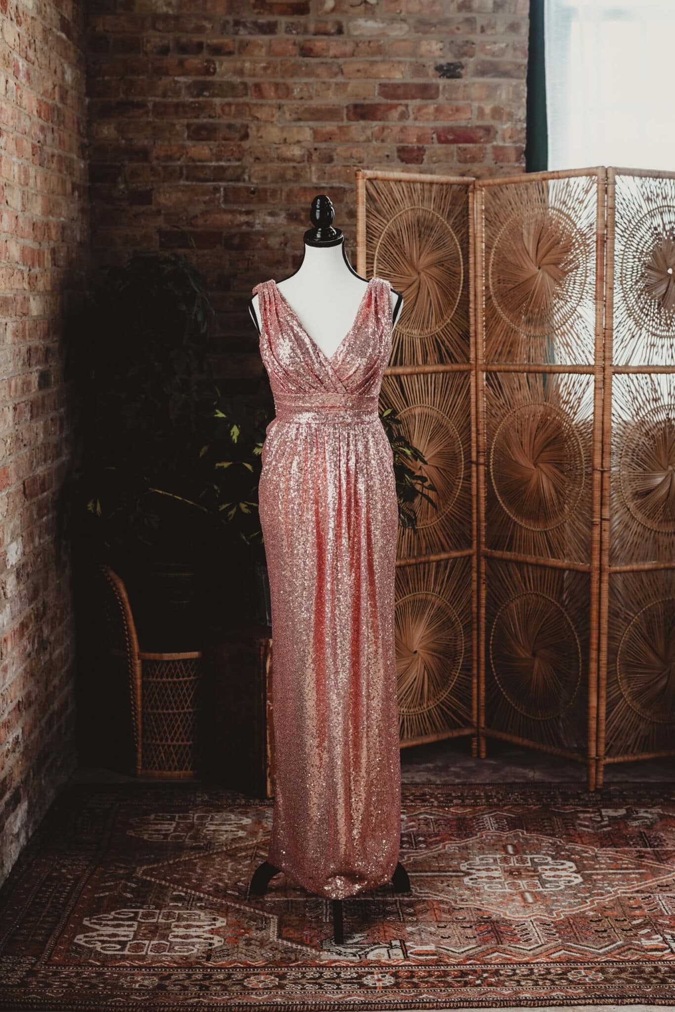 A shimmering pink sequin dress displayed on a black mannequin against a rustic brick wall and ornate rattan room divider