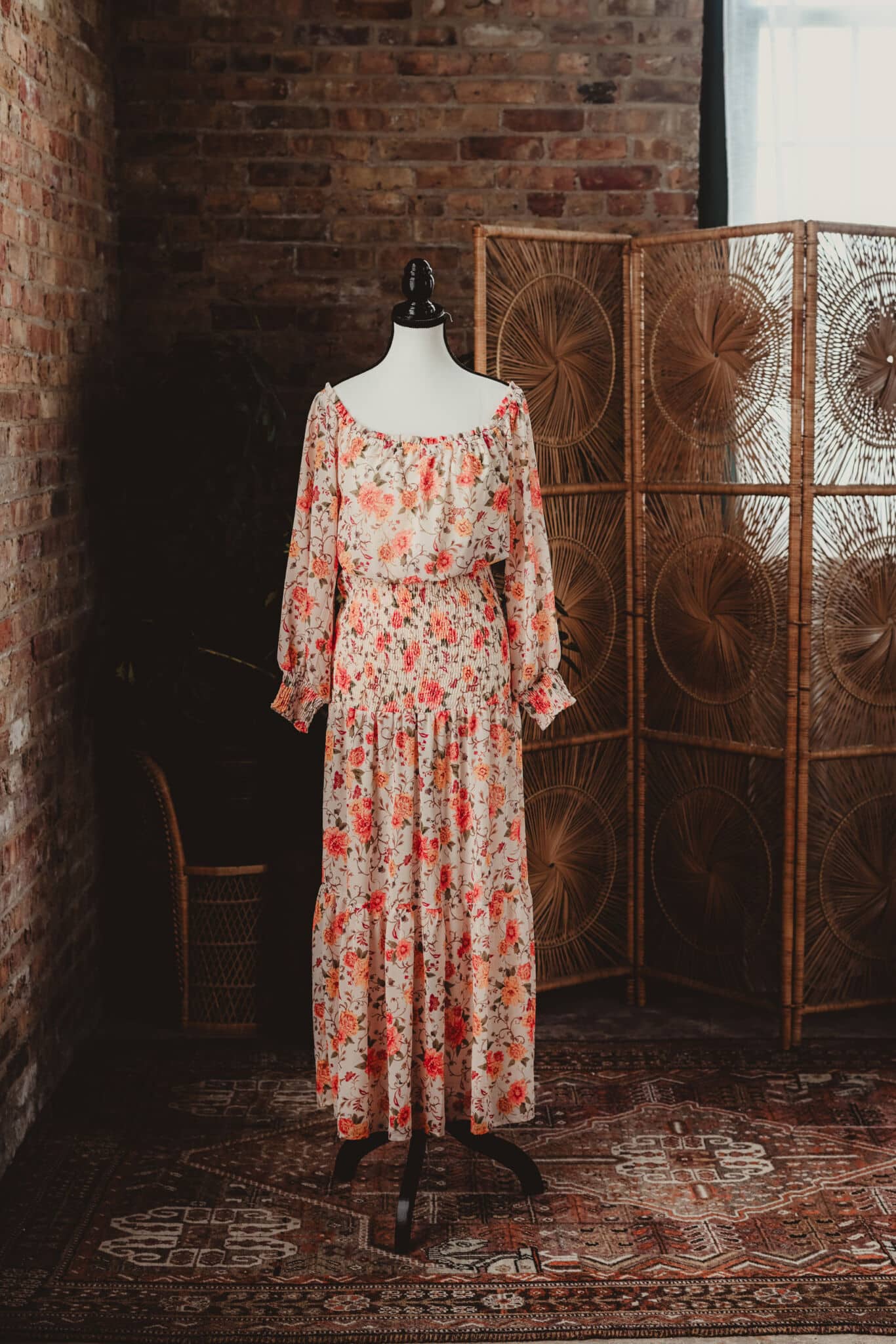 A floral dress with red and pink roses on a cream fabric displayed on a mannequin, set against a rustic brick backdrop and woven room dividers.