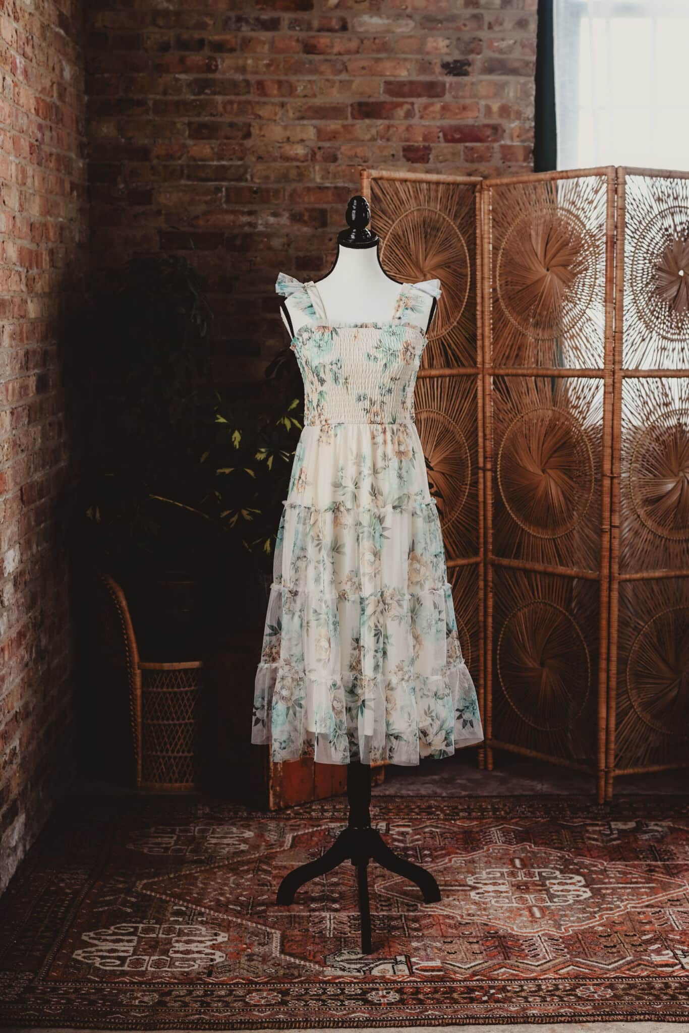 Vintage-inspired floral dress on a mannequin in a rustic setting with a brick wall and decorative rattan screen