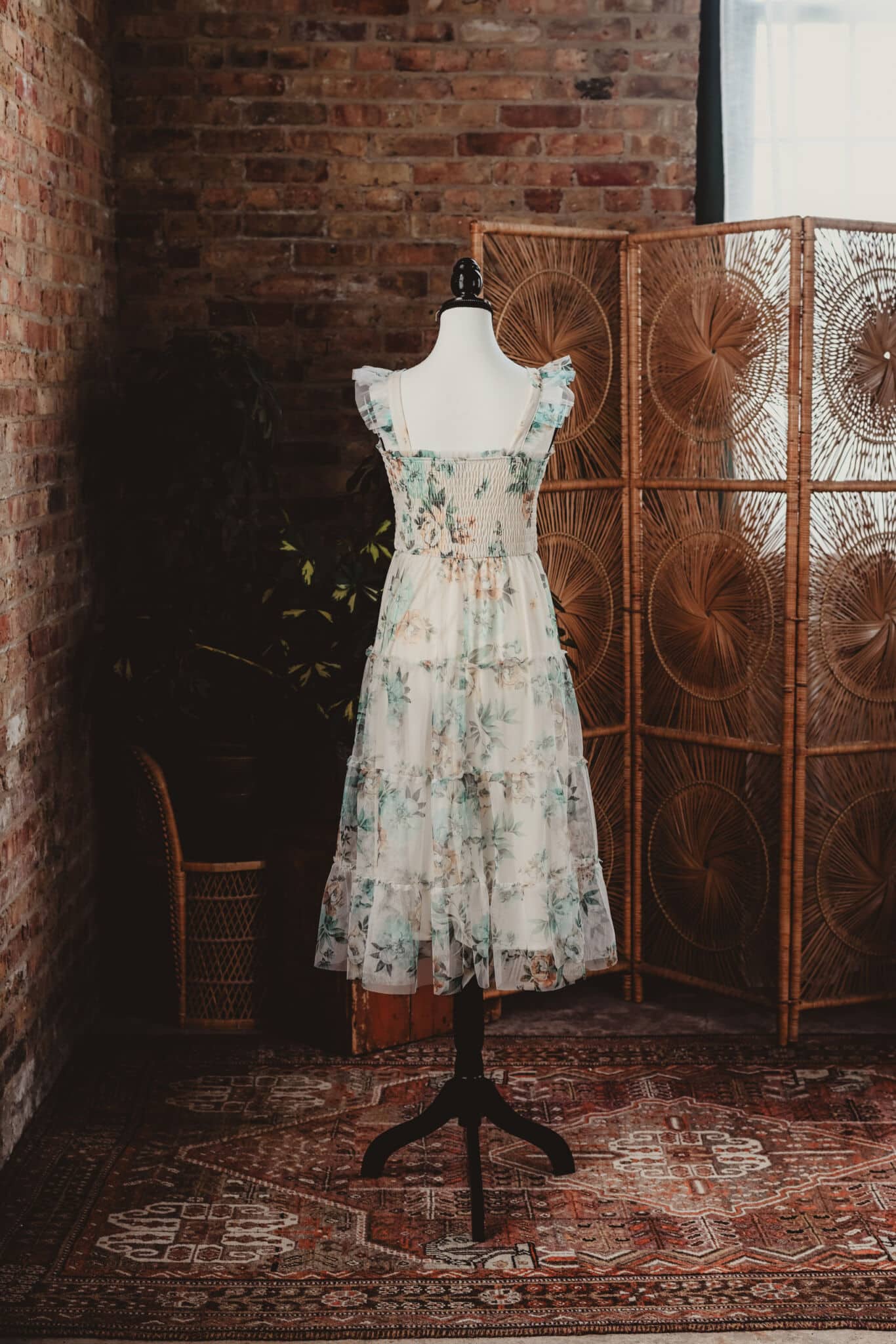 A mannequin displaying a vintage floral dress in a rustic room with brick walls, a patterned rug, and a wicker screen