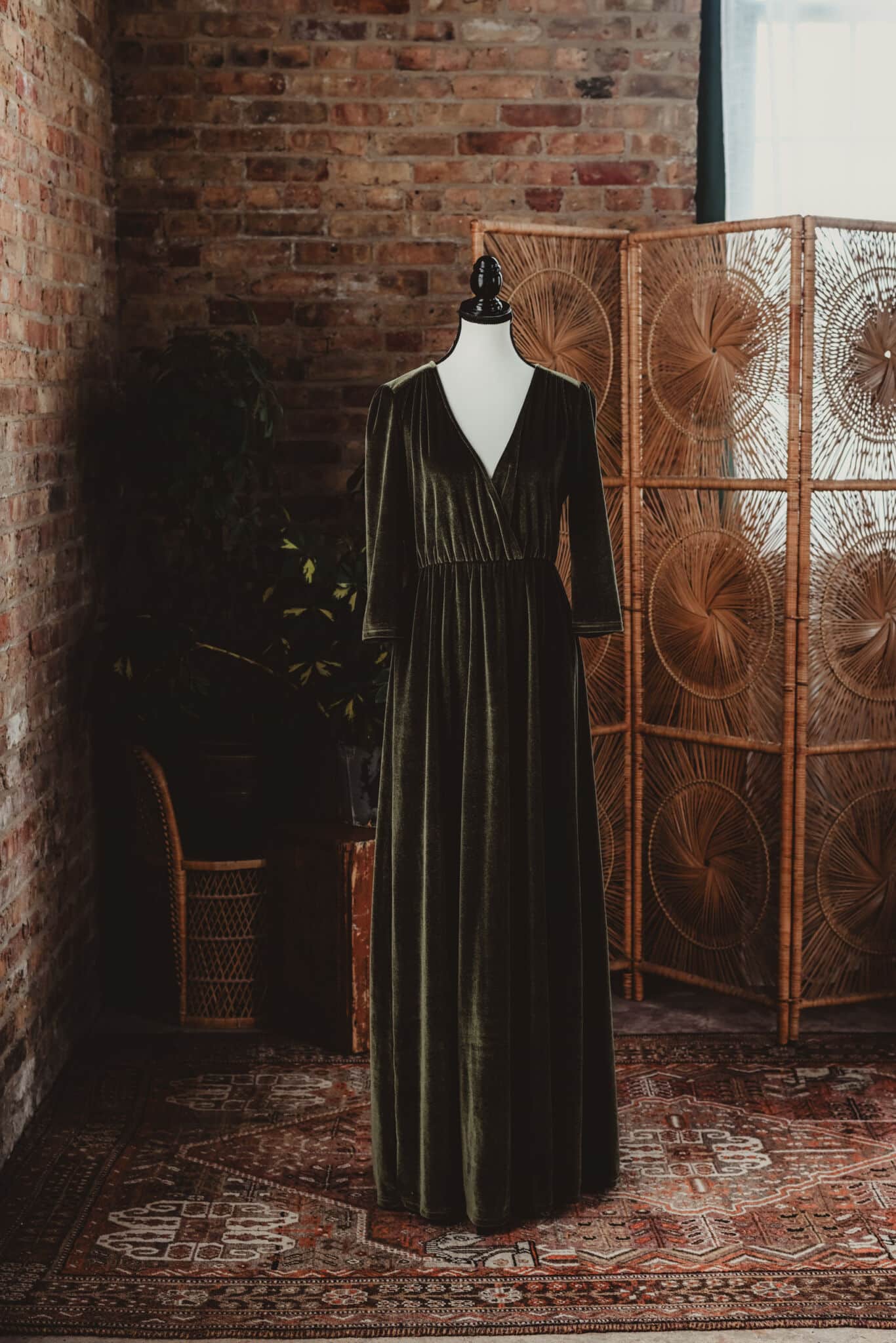 A vintage deep green velvet dress displayed on a mannequin against a rustic brick wall backdrop, accompanied by wicker furniture and lush indoor plants.