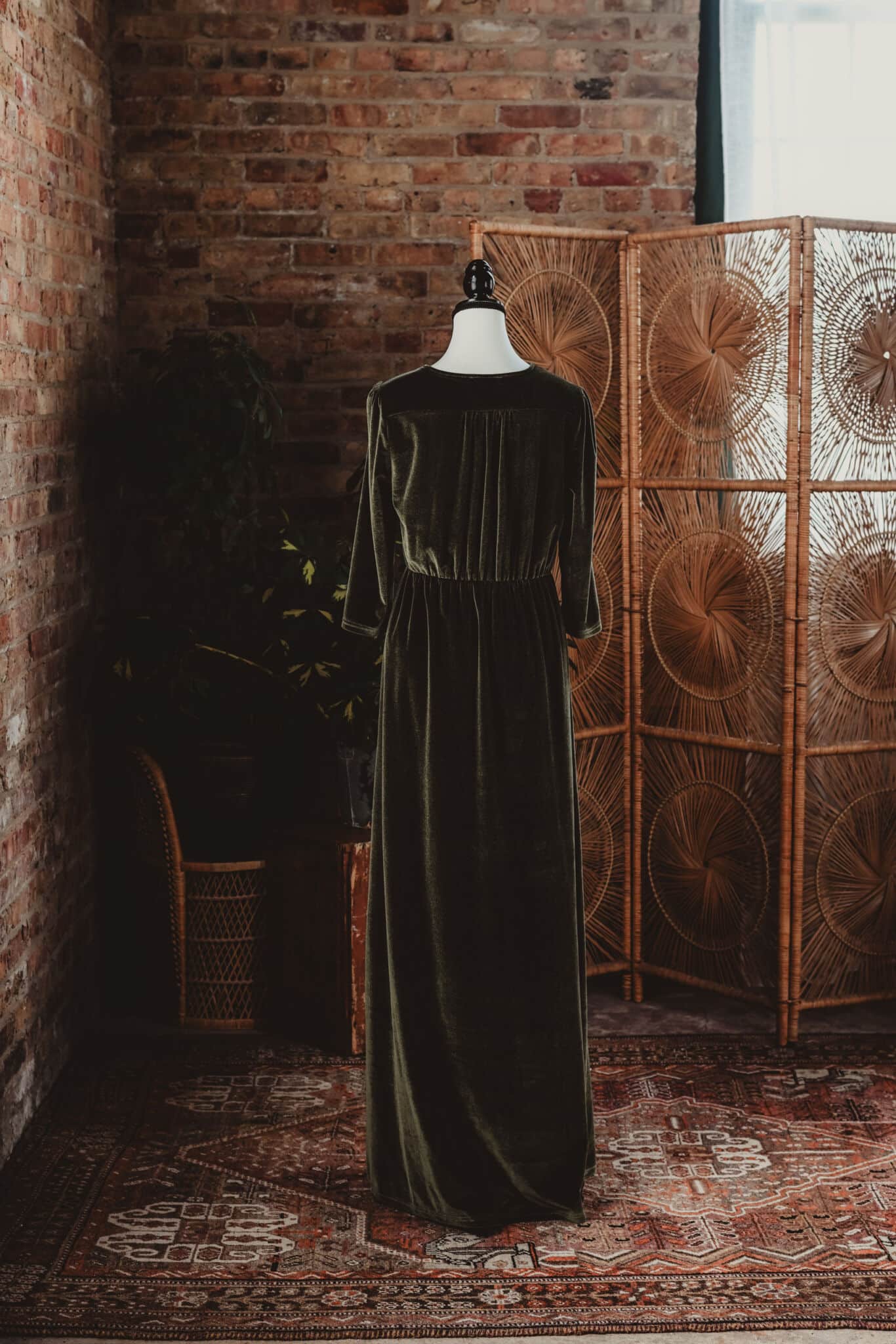 A vintage green velvet dress on a mannequin positioned against an exposed brick wall, with a rattan room divider and rustic carpet in an artistically lit setting