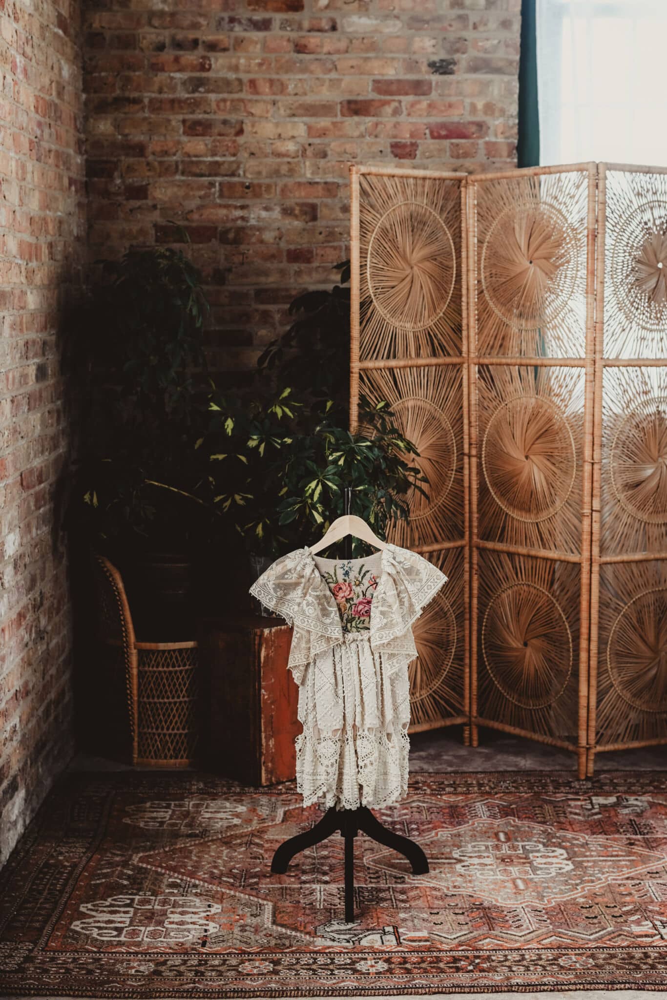 An elegant vintage lace dress with floral embroidery displayed on a stand against a rustic brick wall, behind a decorative wooden screen and surrounded by lush plants.