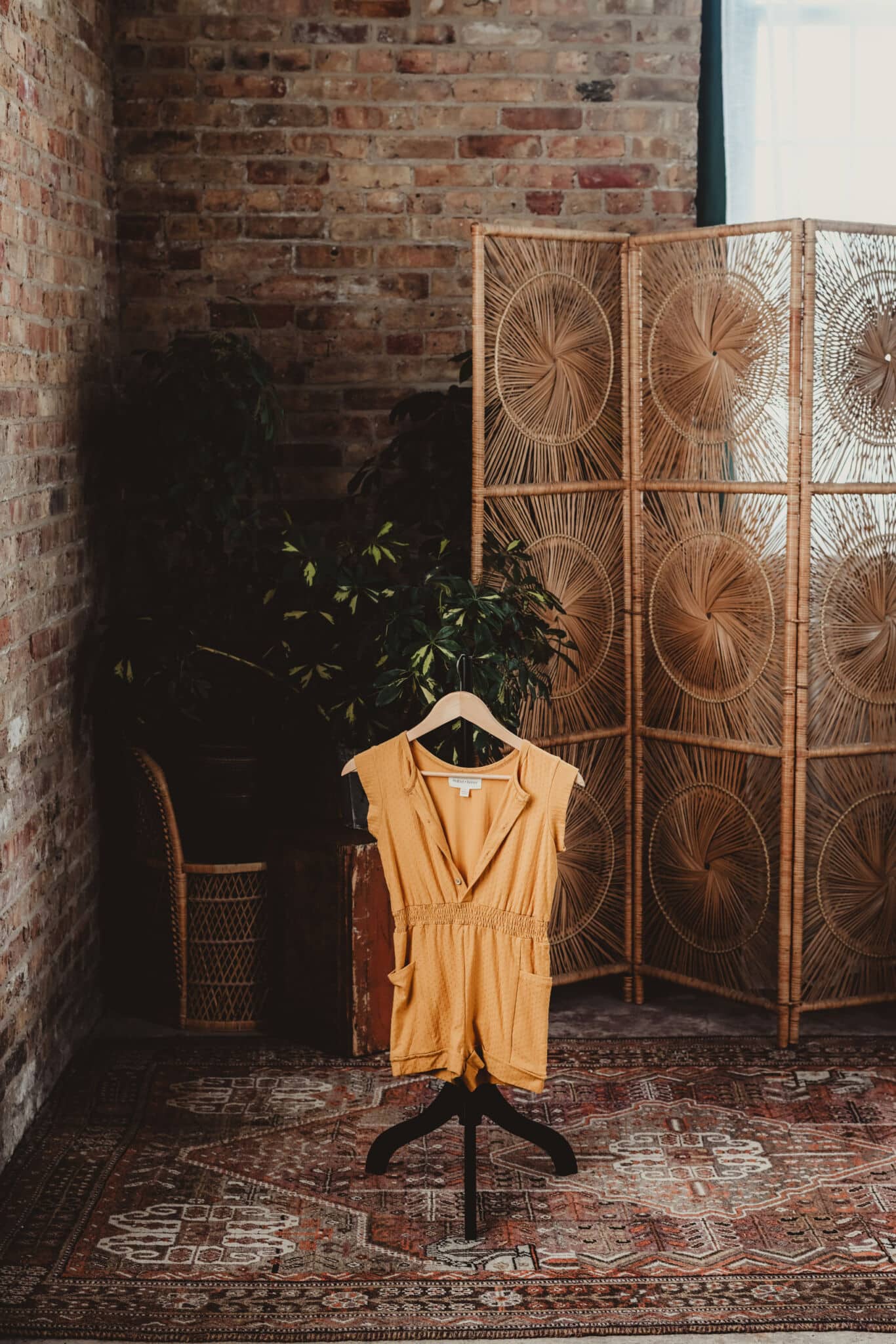 A vibrant yellow vintage dress displayed on a mannequin in a rustic room with a brick wall, ornate carpet, and a wicker room divider.