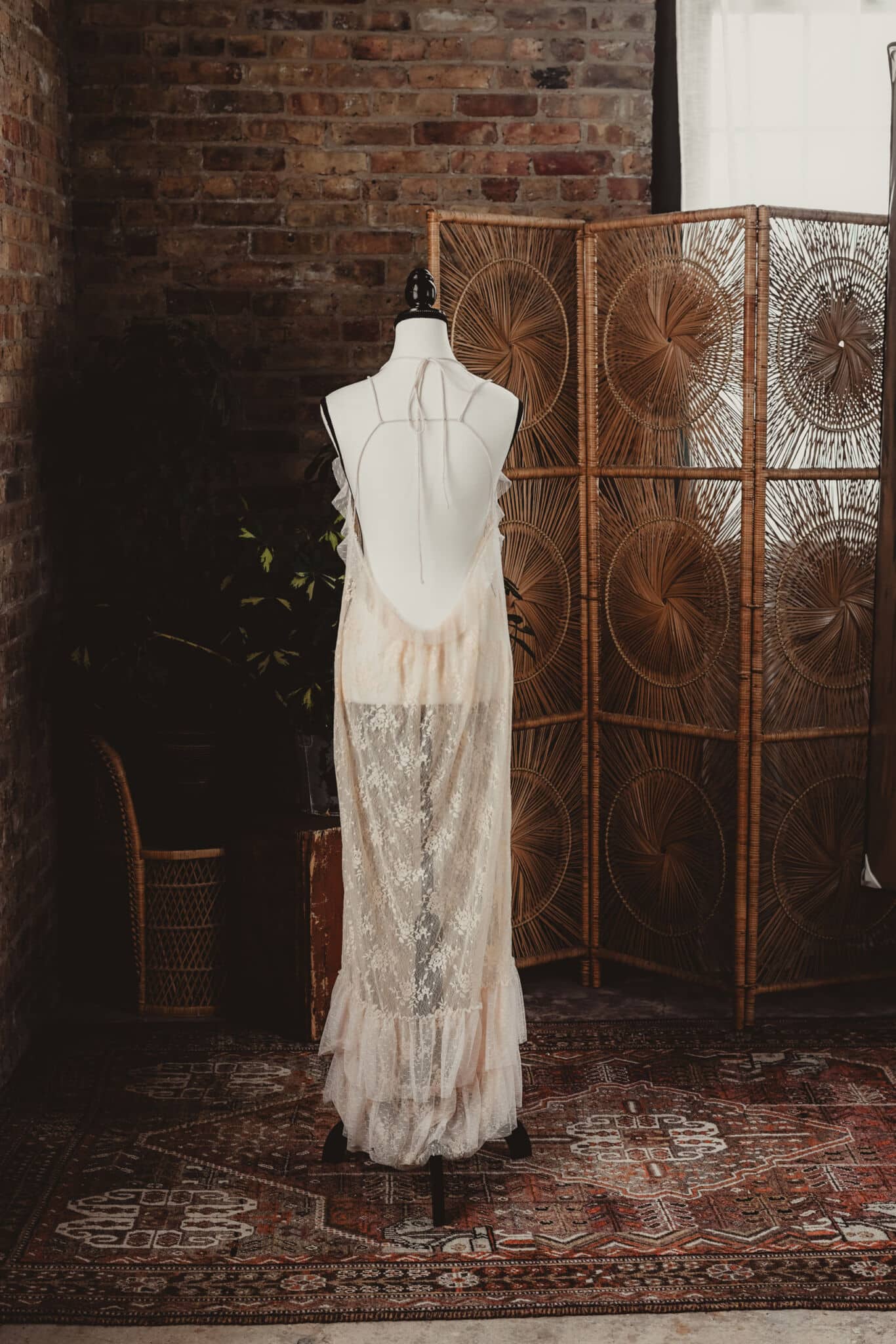 Vintage lace wedding dress on a mannequin in a bohemian styled room with a rattan screen and brick wall