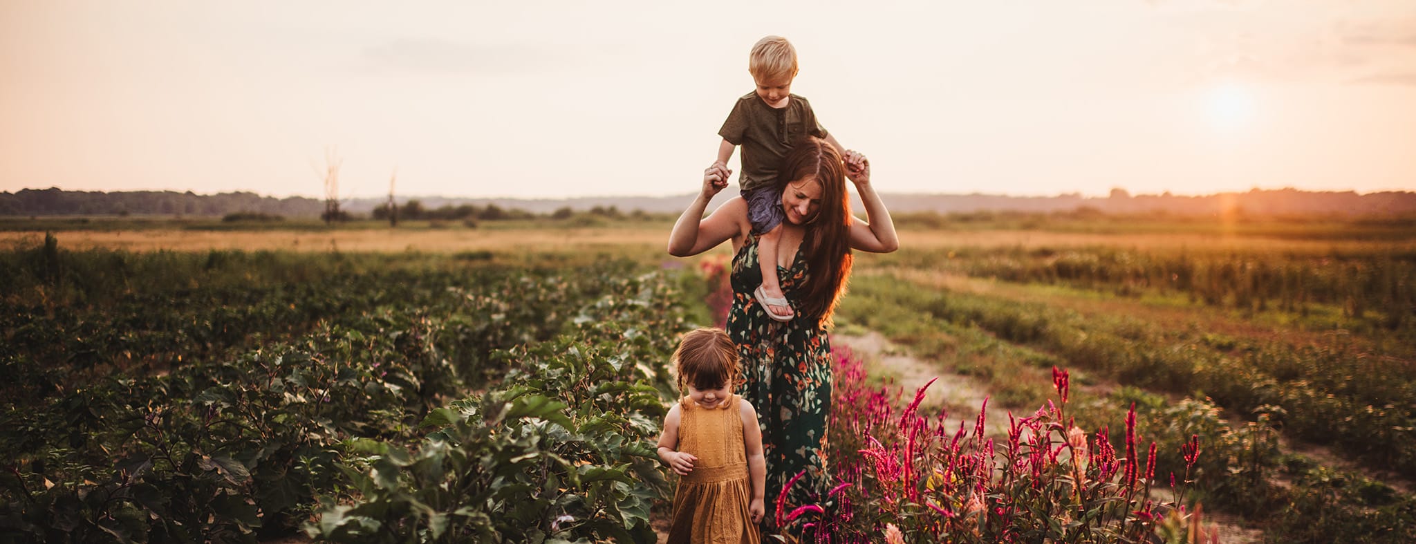 A woman carrying a young boy on her shoulders, with another child walking beside them in a vibrant flower field at sunset.