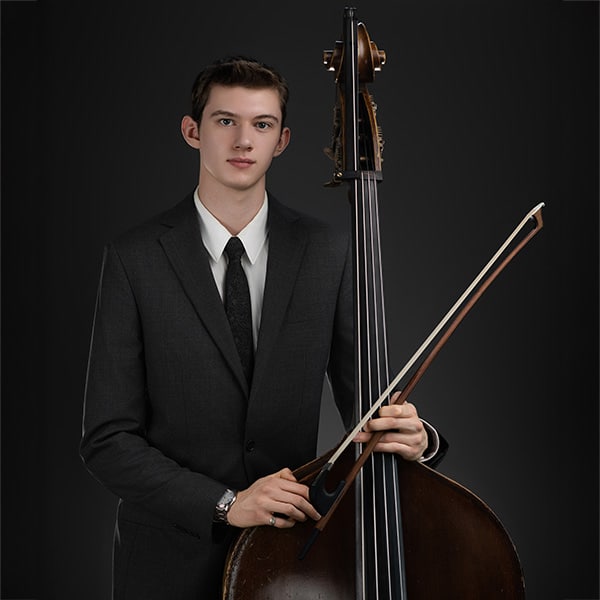 young_male_musician_playing_double_bass A young man in a formal suit holding a double bass and bow in a studio setting against a dark background