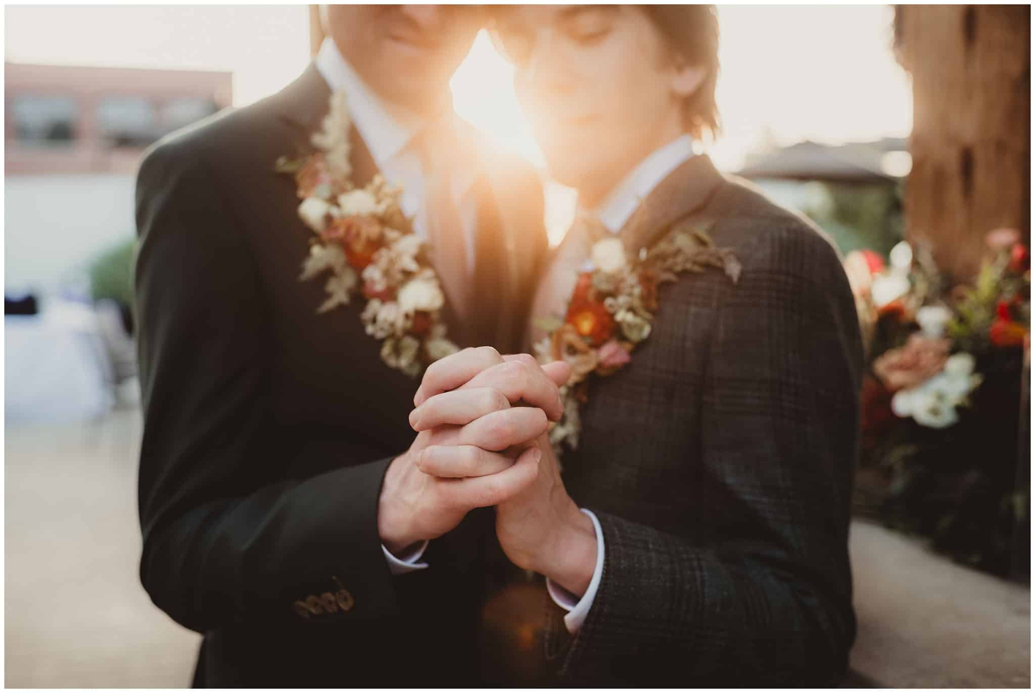 Two grooms in suits with floral corsages holding hands during a sunset at their wedding