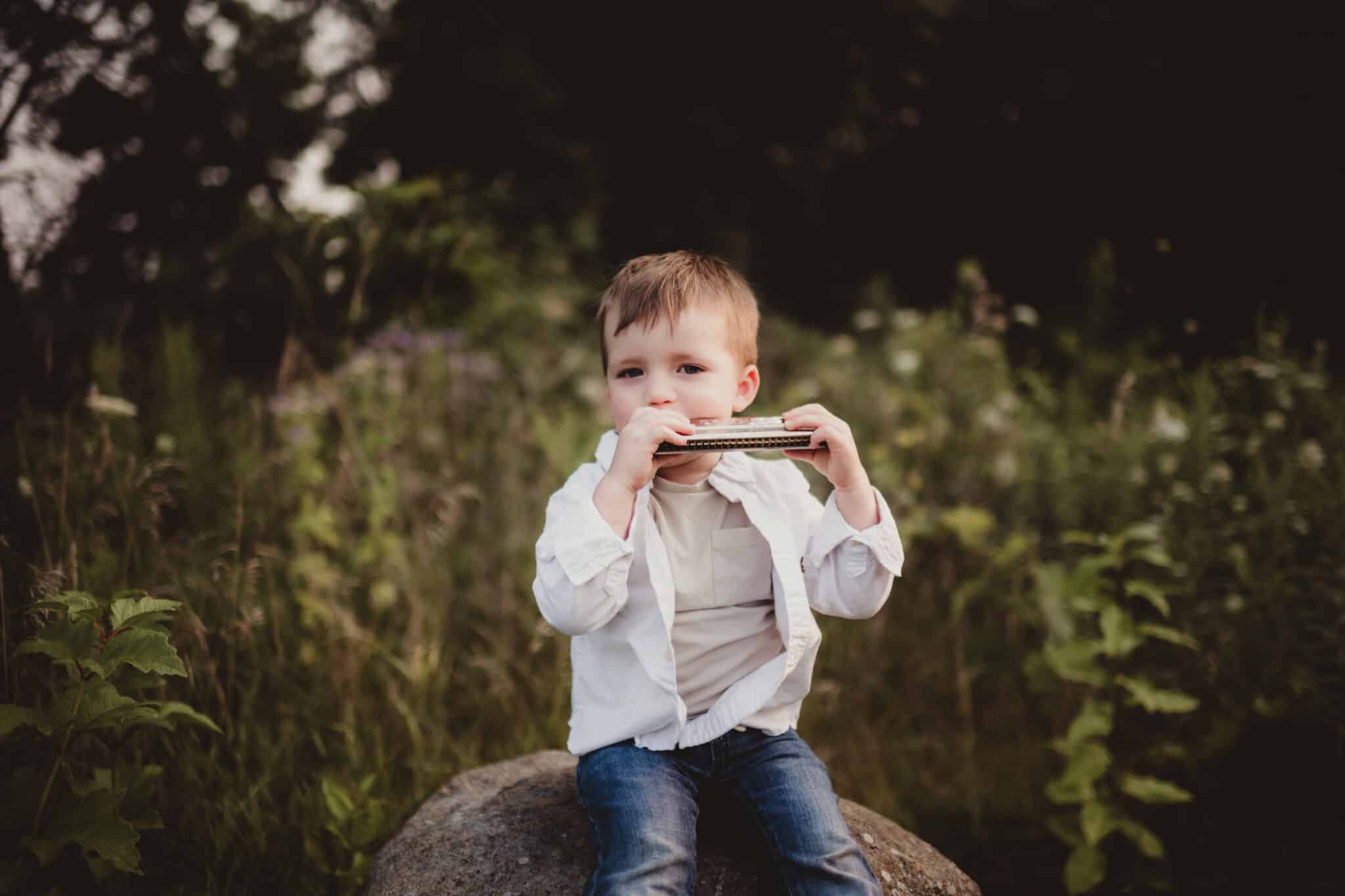 Young boy sitting on a rock playing a harmonica in a natural setting with soft focus background