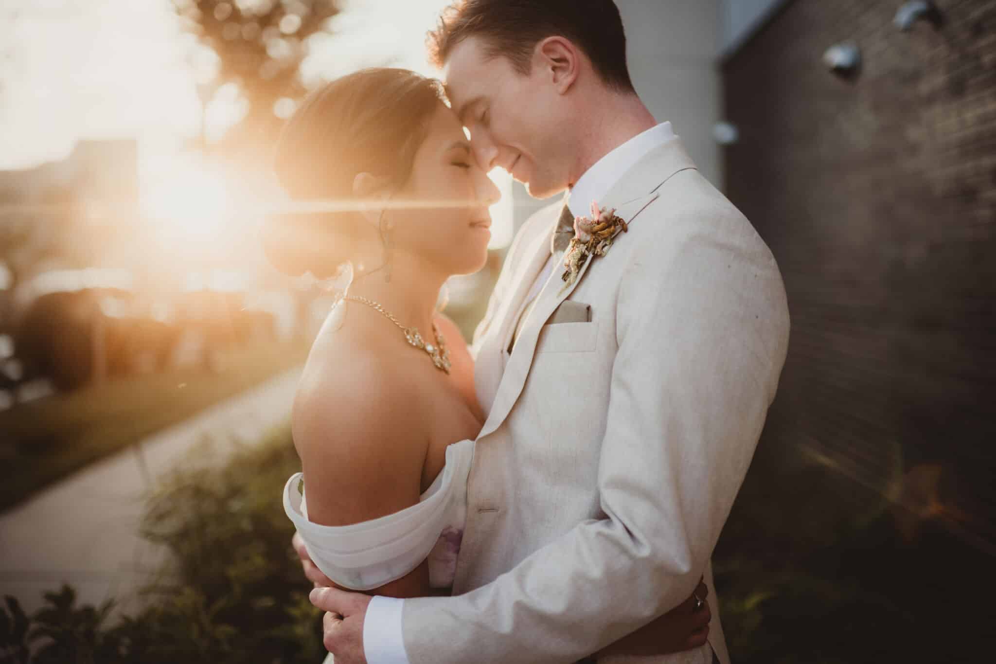 Bride and groom embracing in a tender moment with the sunset behind them, creating a warm glow around them.