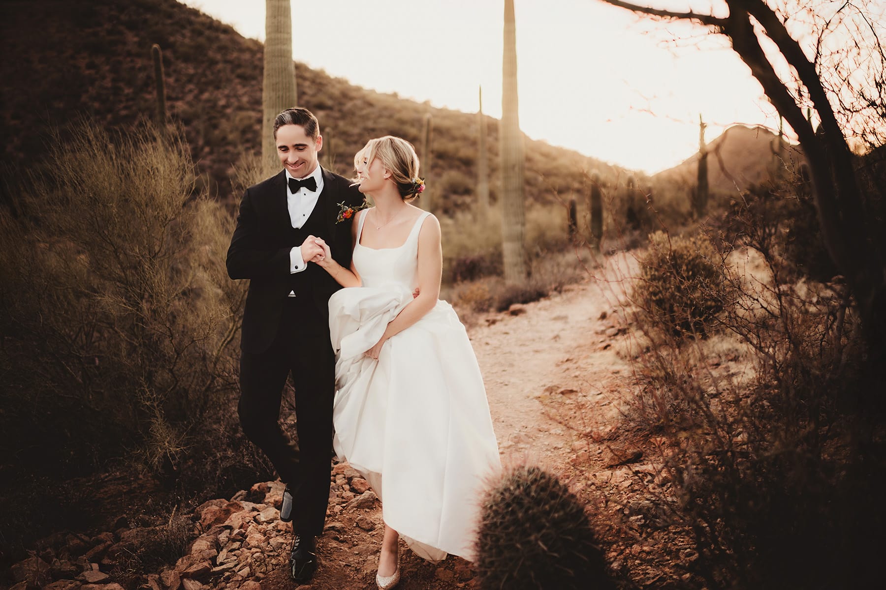 Wedding couple walking hand in hand in a desert landscape at sunset, surrounded by towering cacti
