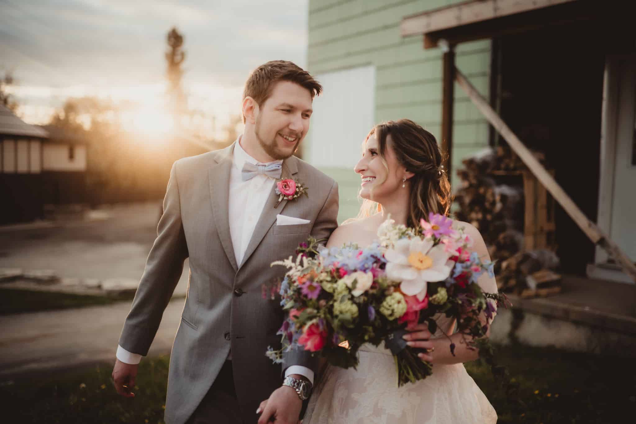 A joyful bride and groom walking hand in hand during sunset, with the bride holding a colorful bouquet of flowers