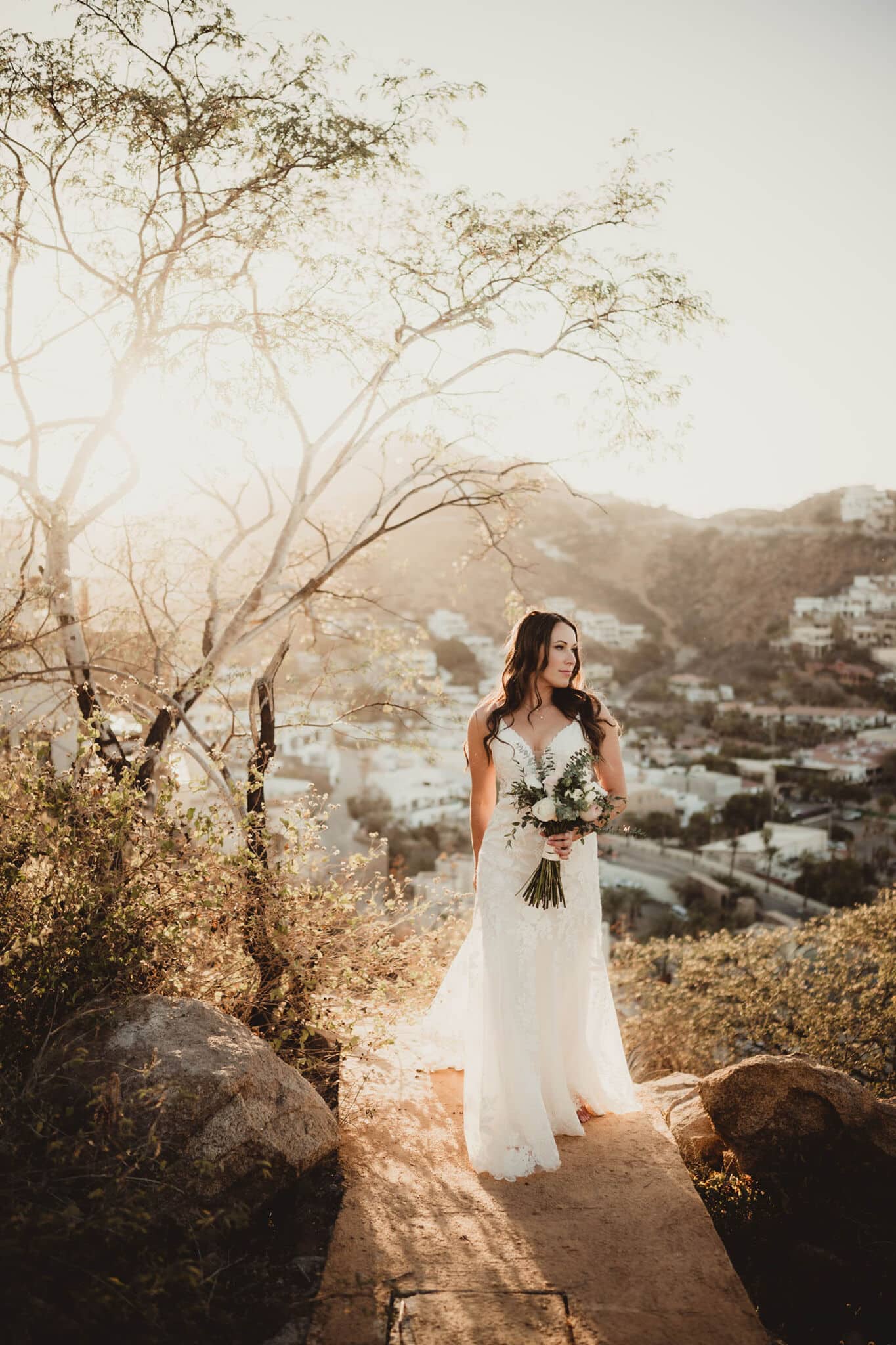 A bride in a flowing white gown holding a bouquet, standing on a hillside path with a sunlit cityscape in the background